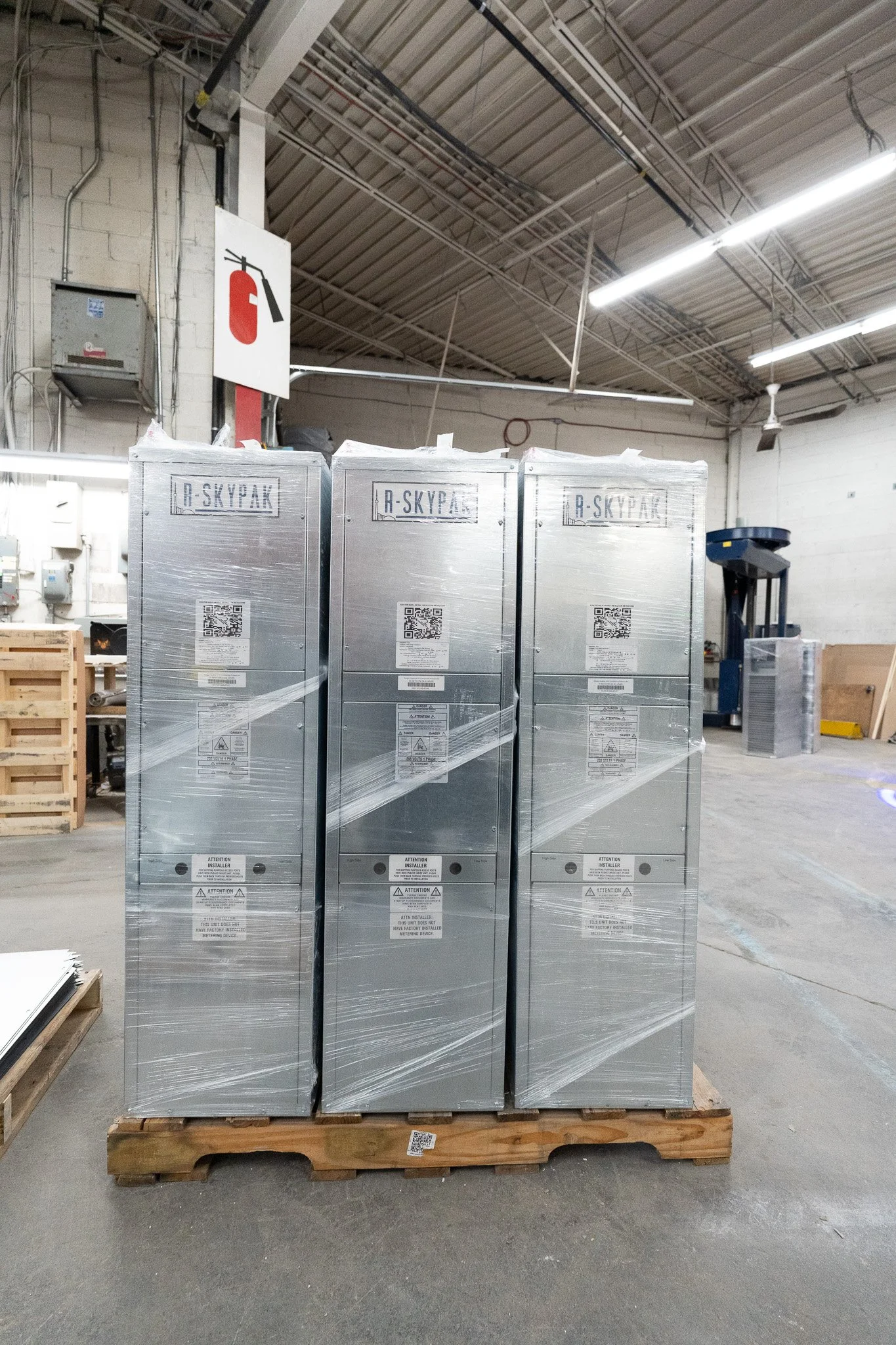 Three large boxed items wrapped in plastic on a wooden pallet inside a warehouse, with a fire extinguisher sign above.