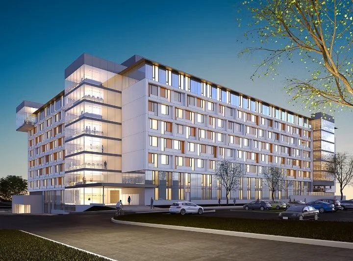 A modern multi-story office building with glass windows and a parking lot in front, during twilight with a clear sky and some trees nearby.