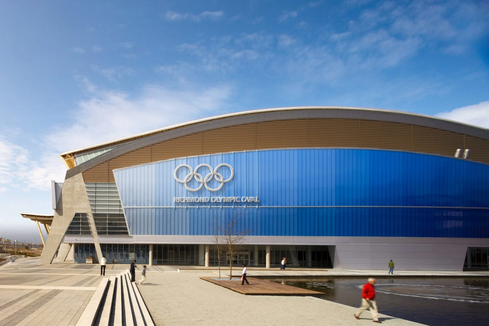 The Richmond Olympic Oval building with the Olympic rings logo and the words 'Richmond Olympic Oval' on a blue facade, with a paved outdoor area and a small water feature in the foreground, under a partly cloudy sky.