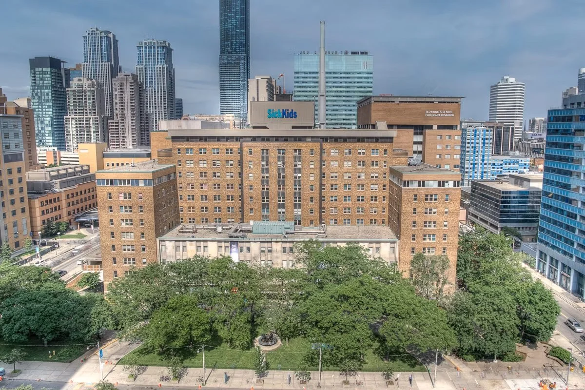 A cityscape with tall modern buildings in the background and an older brick building in the foreground surrounded by green trees.