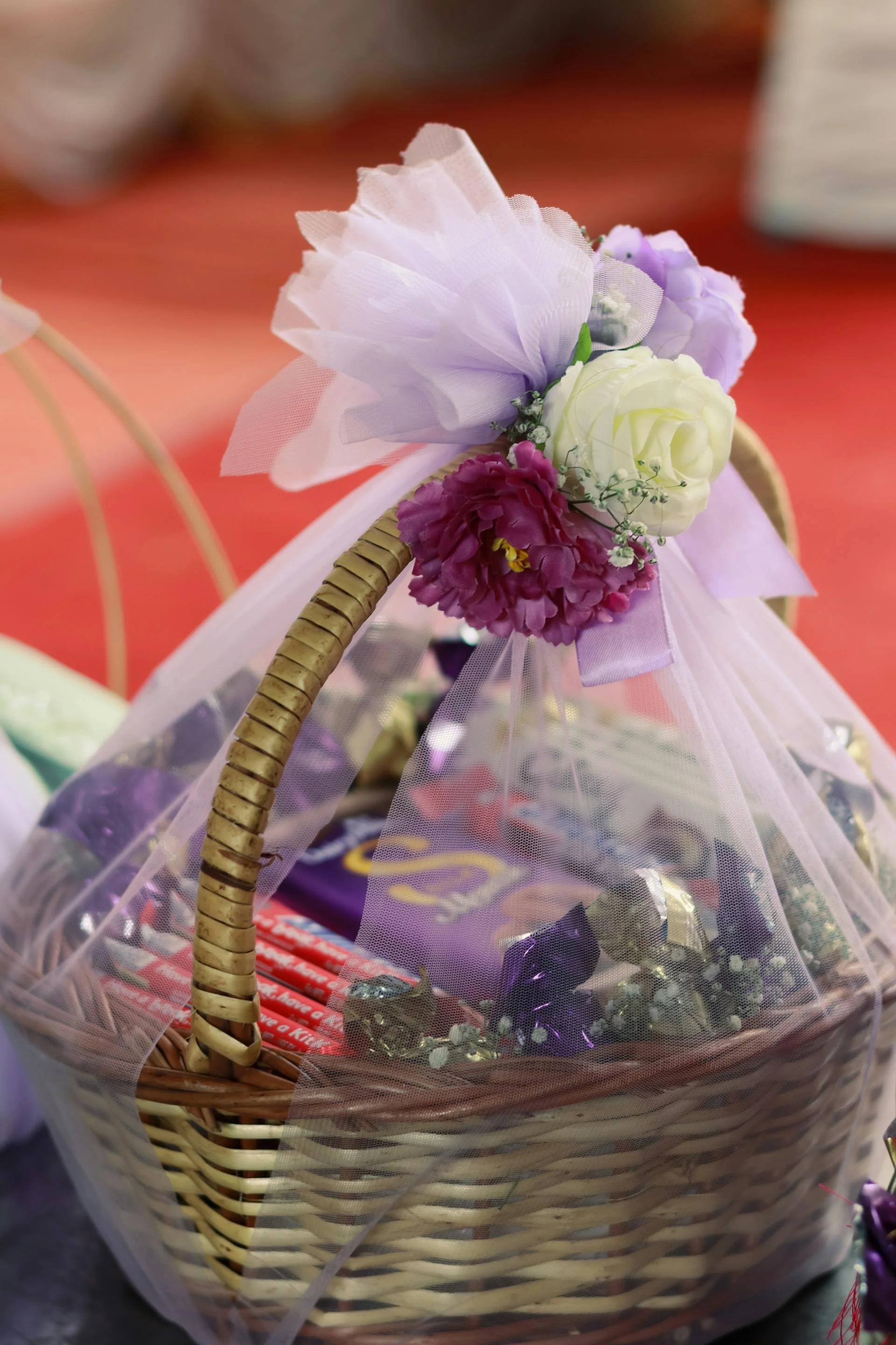 A decorated gift basket with a pink bow and artificial flowers including white and purple roses and purple and pink flowers, wrapped in clear plastic.