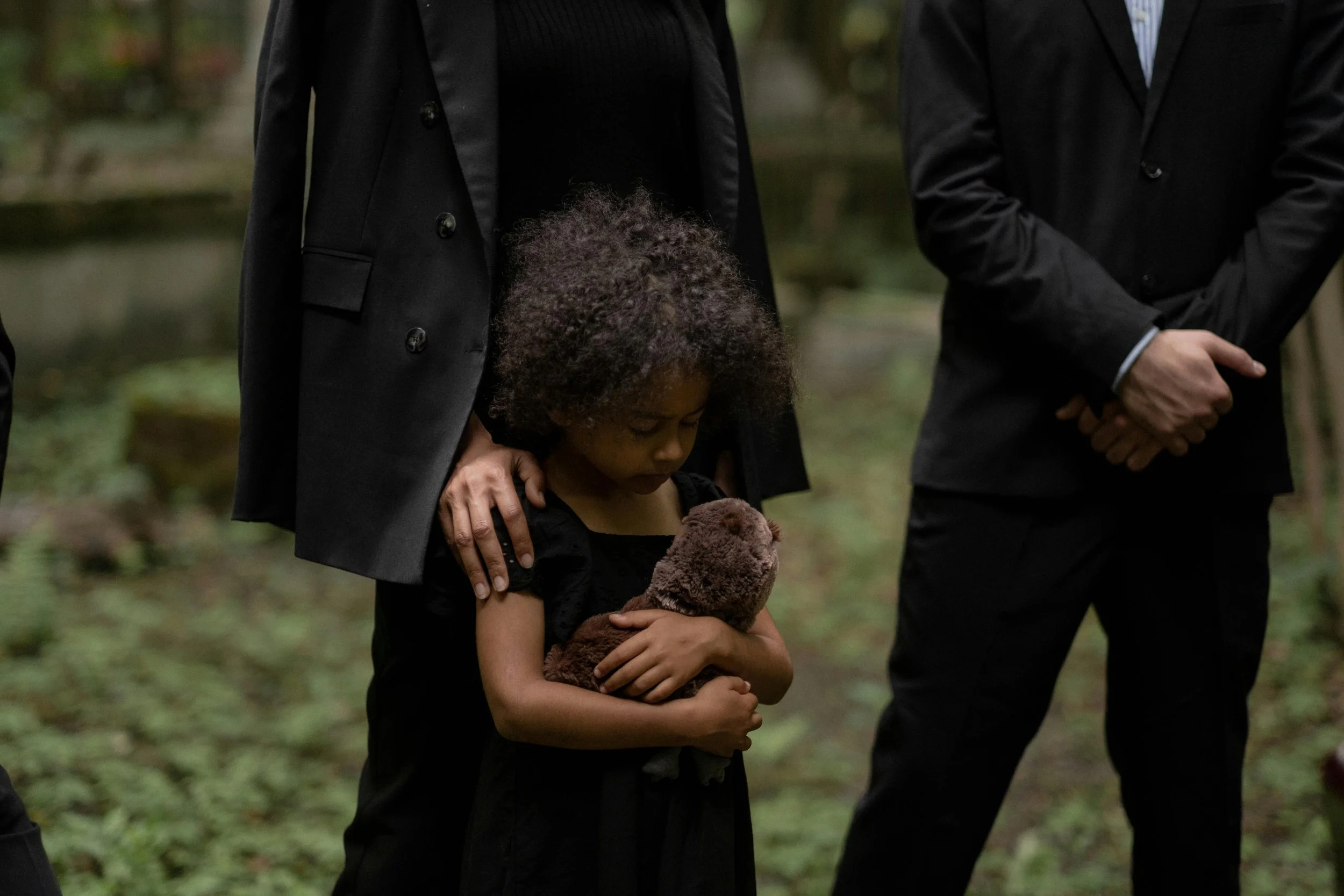 A young girl holding a brown teddy bear, with her eyes closed, standing between two adults in formal black suits, outdoors in a natural setting.