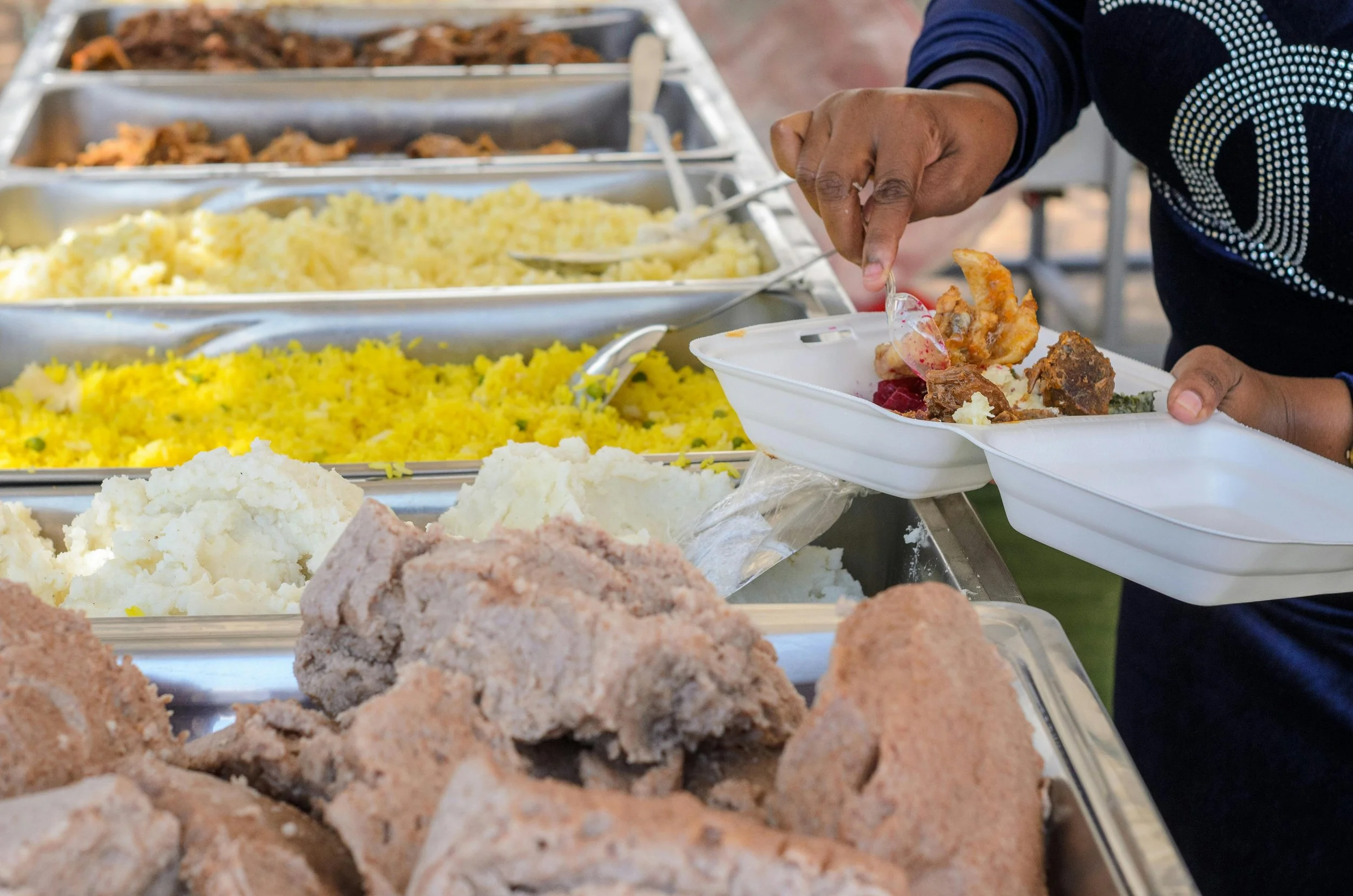 Person serving food at a buffet with various dishes including mashed potatoes, yellow rice, and fried meats.