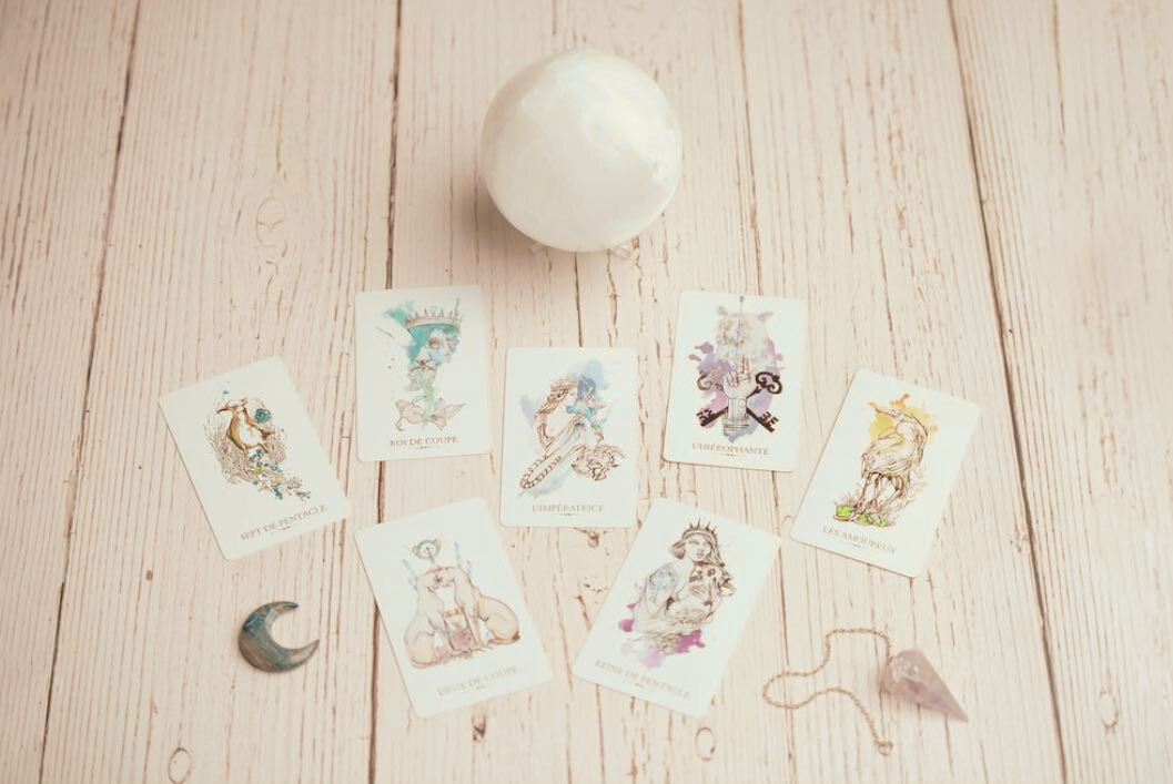 Cartes de tarot, lune en bois, pierre en pendentif, boule de cristal sur un plan en bois clair.