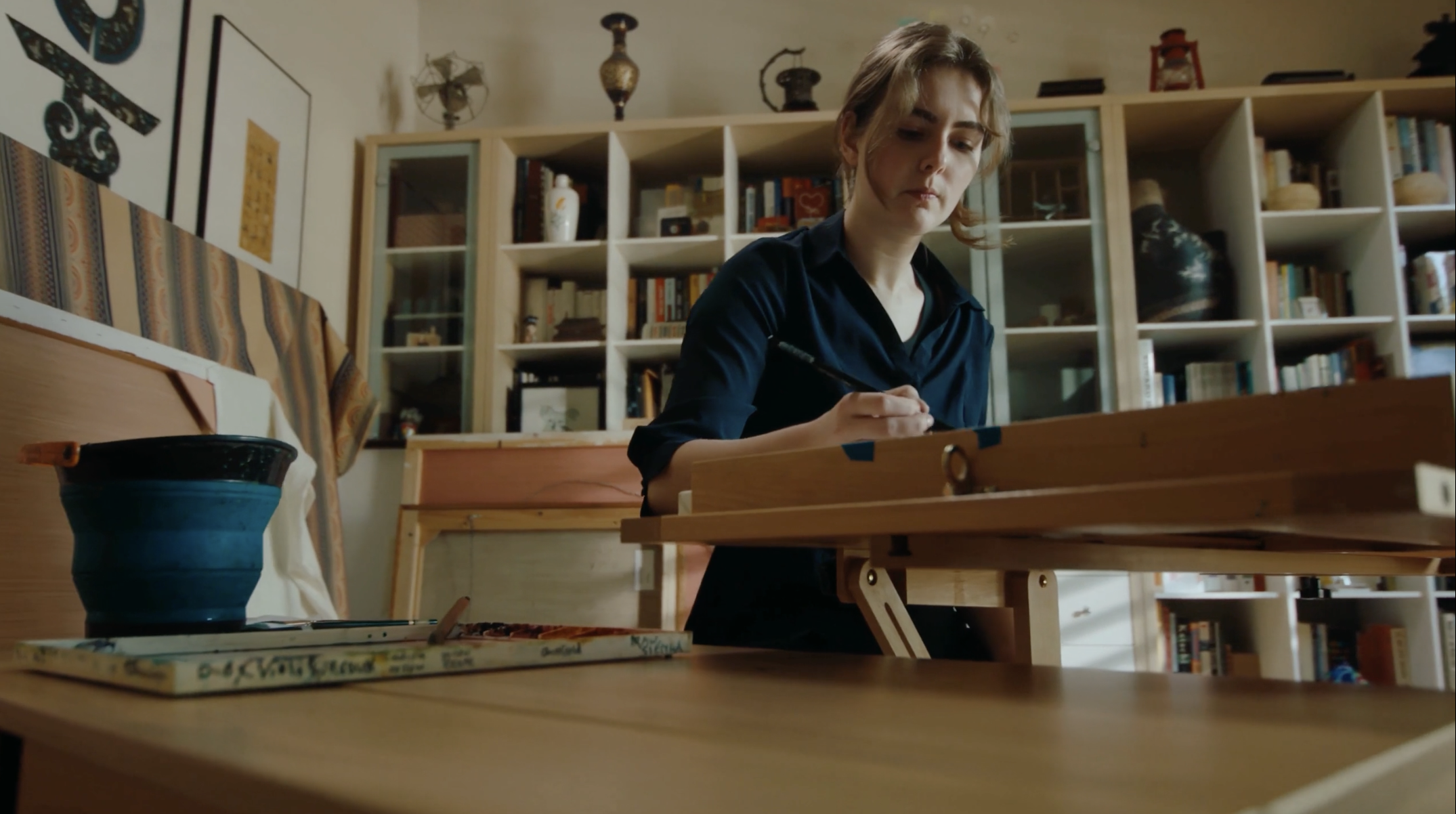 A woman with short, wavy hair, wearing a navy blue top, is sitting at a wooden table, writing or drawing on a piece of paper.