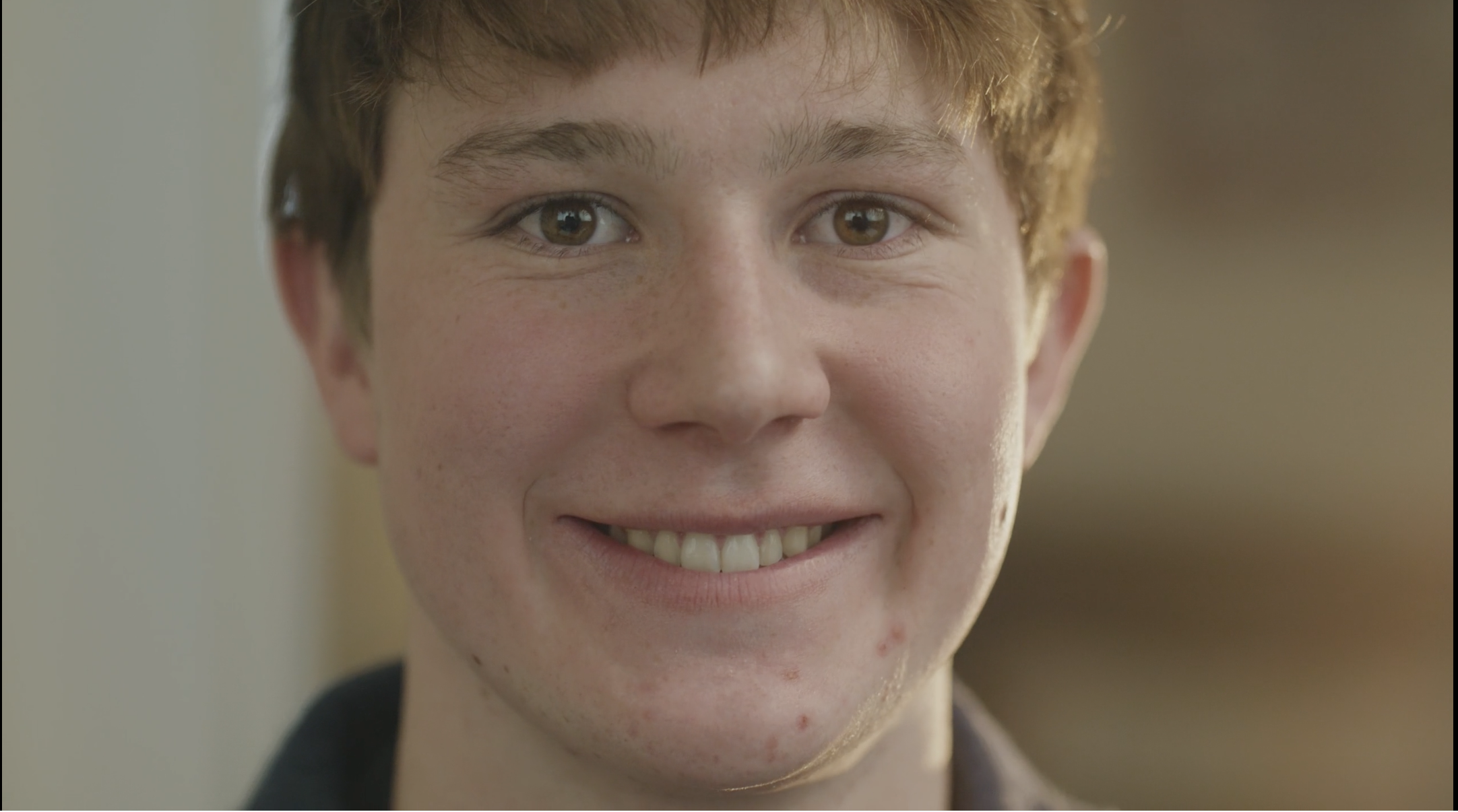Close-up of a young person's smiling face, showing brown eyes, freckles, and short hair, against a neutral background.