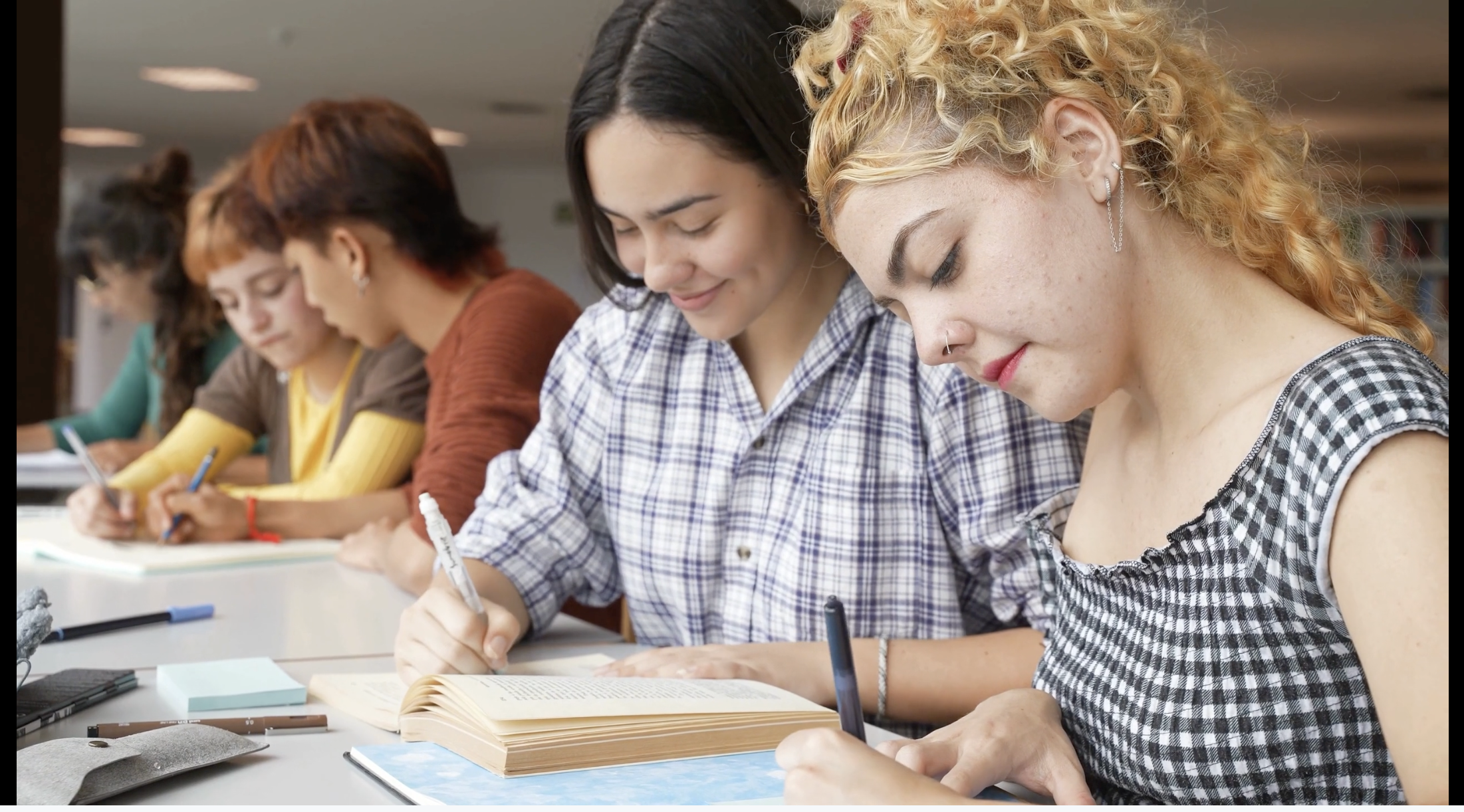 A group of diverse young women sitting at a table, studying or taking notes, with books and writing materials in front of them, in a well-lit indoor setting.