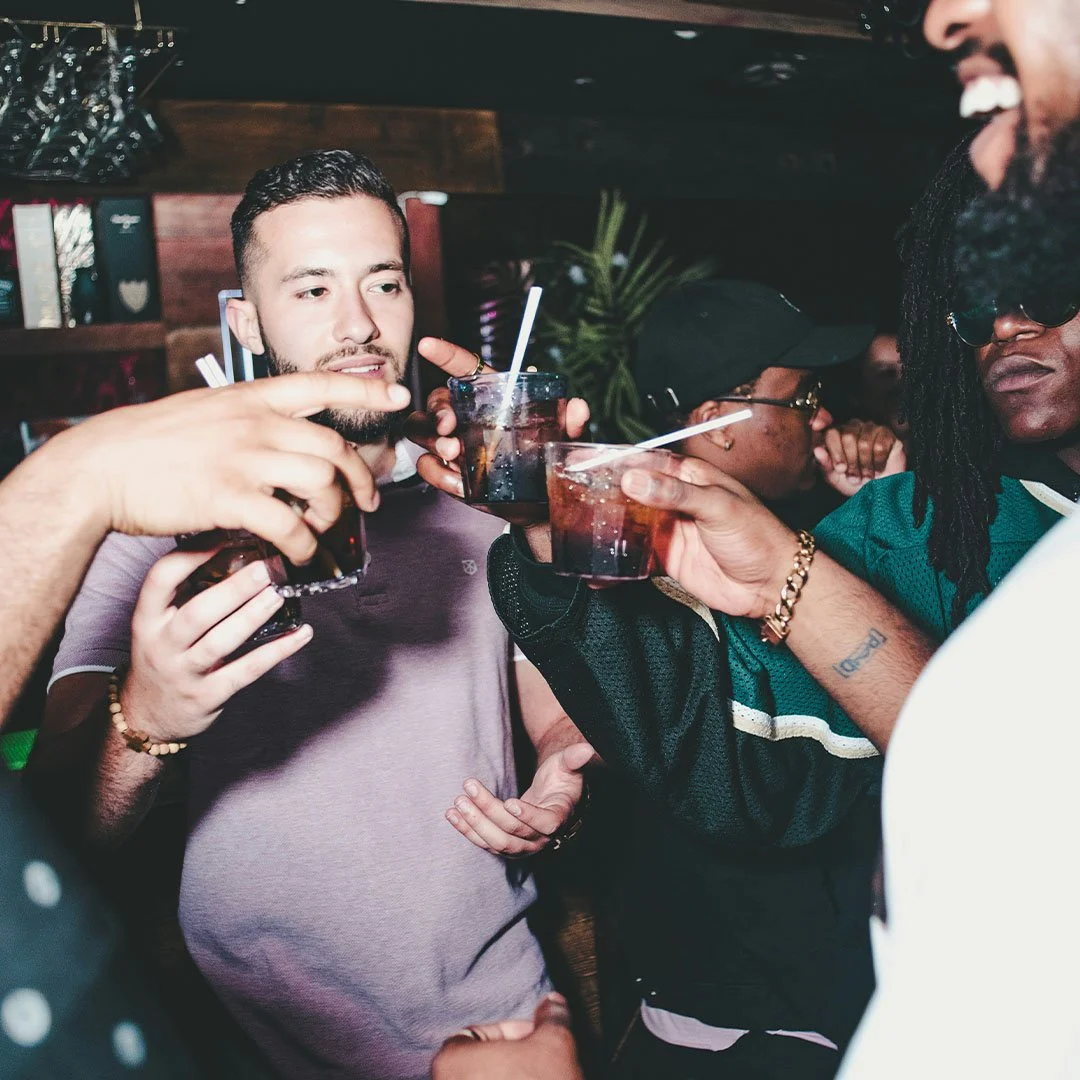 A group of friends toasting drinks at a party or bar, smiling and enjoying each other's company.