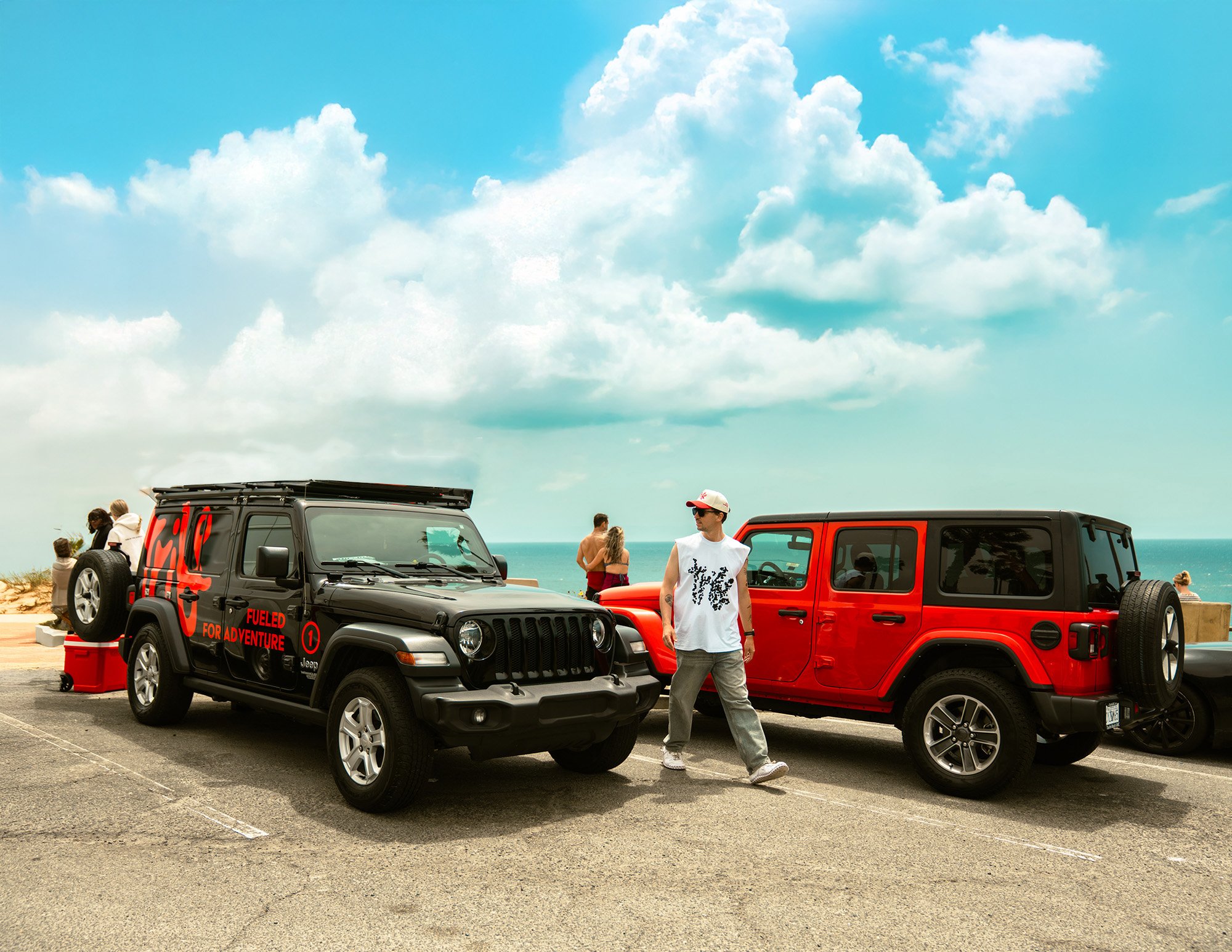 Two Jeep vehicles parked at a beachside parking lot with people near the ocean, a man walking between the vehicles, and a blue sky with clouds overhead.