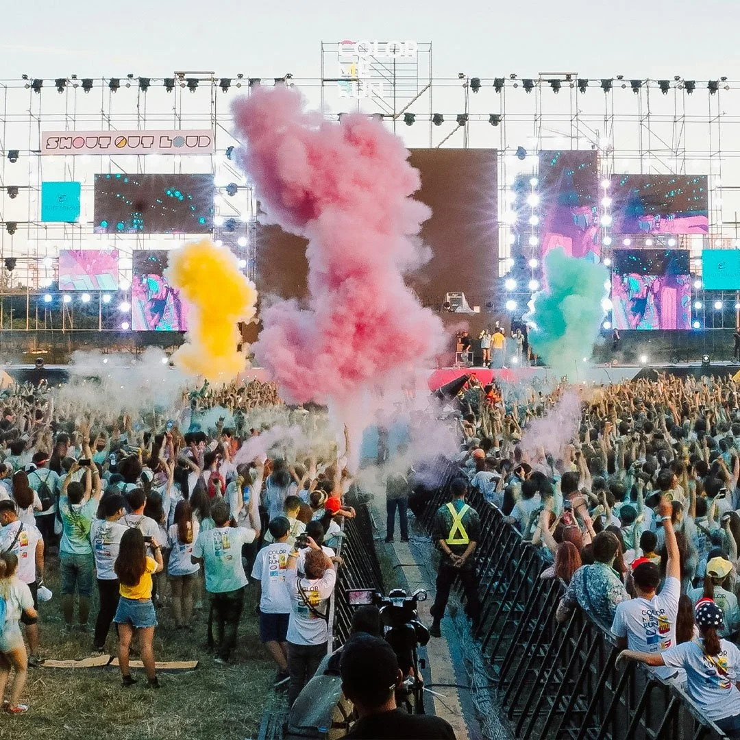 Crowd at a music festival with colorful smoke clouds in yellow, pink, and green in front of a large stage with bright lights and screens.