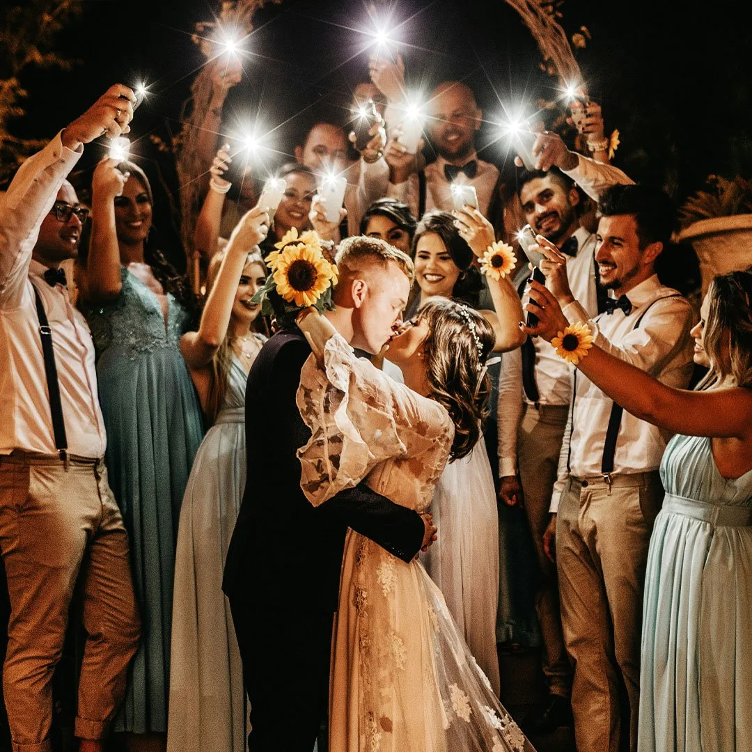 A wedding couple kissing surrounded by friends taking photos with sparklers at night.