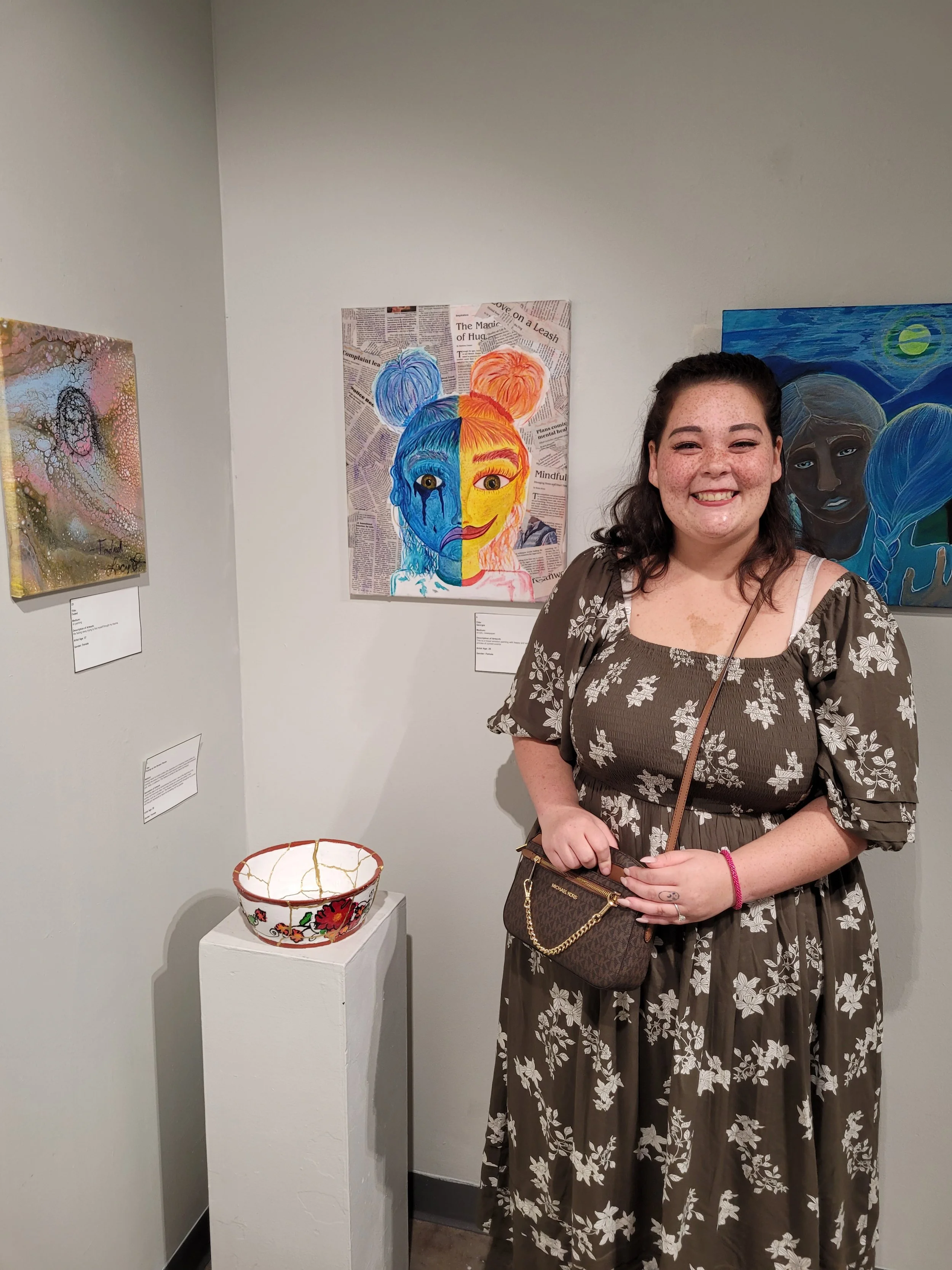 A young woman with curly brown hair and freckles, wearing a brown dress with white floral patterns, stands smiling in an art gallery. Behind her, two colorful paintings of women are displayed on the wall, and a decorative ceramic bowl is on a white pedestal nearby.