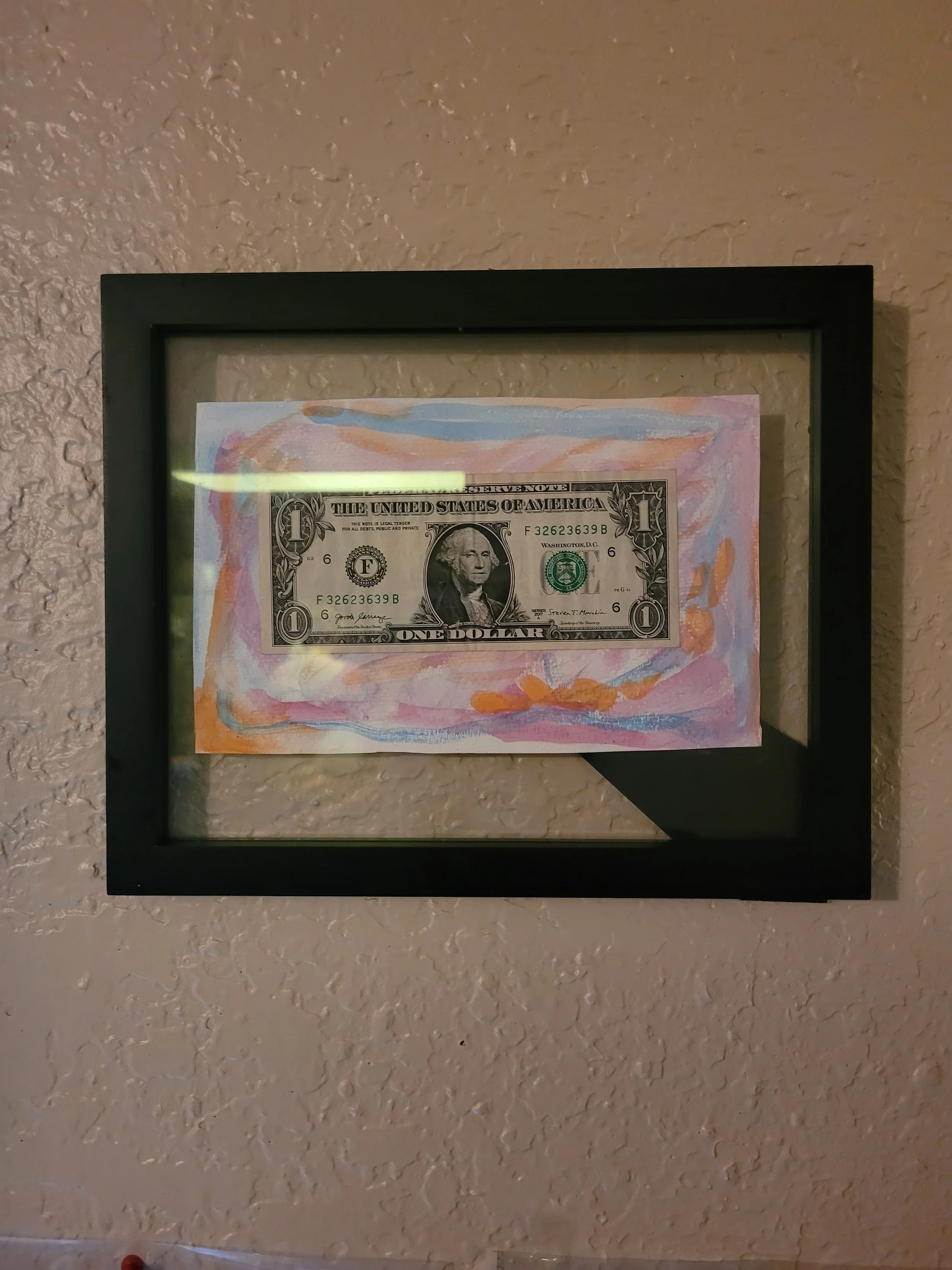 Framed artwork featuring a $1 bill on top of a colorful abstract painting, displayed on a textured beige wall.