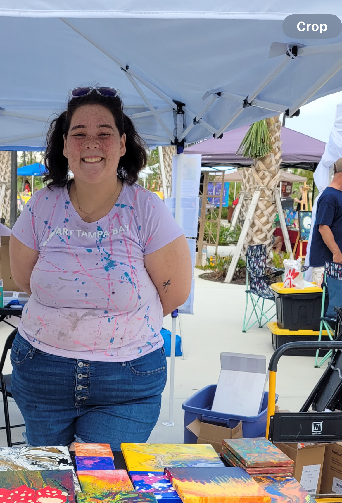 A woman smiling at an outdoor art market, standing behind colorful abstract art paintings on display.