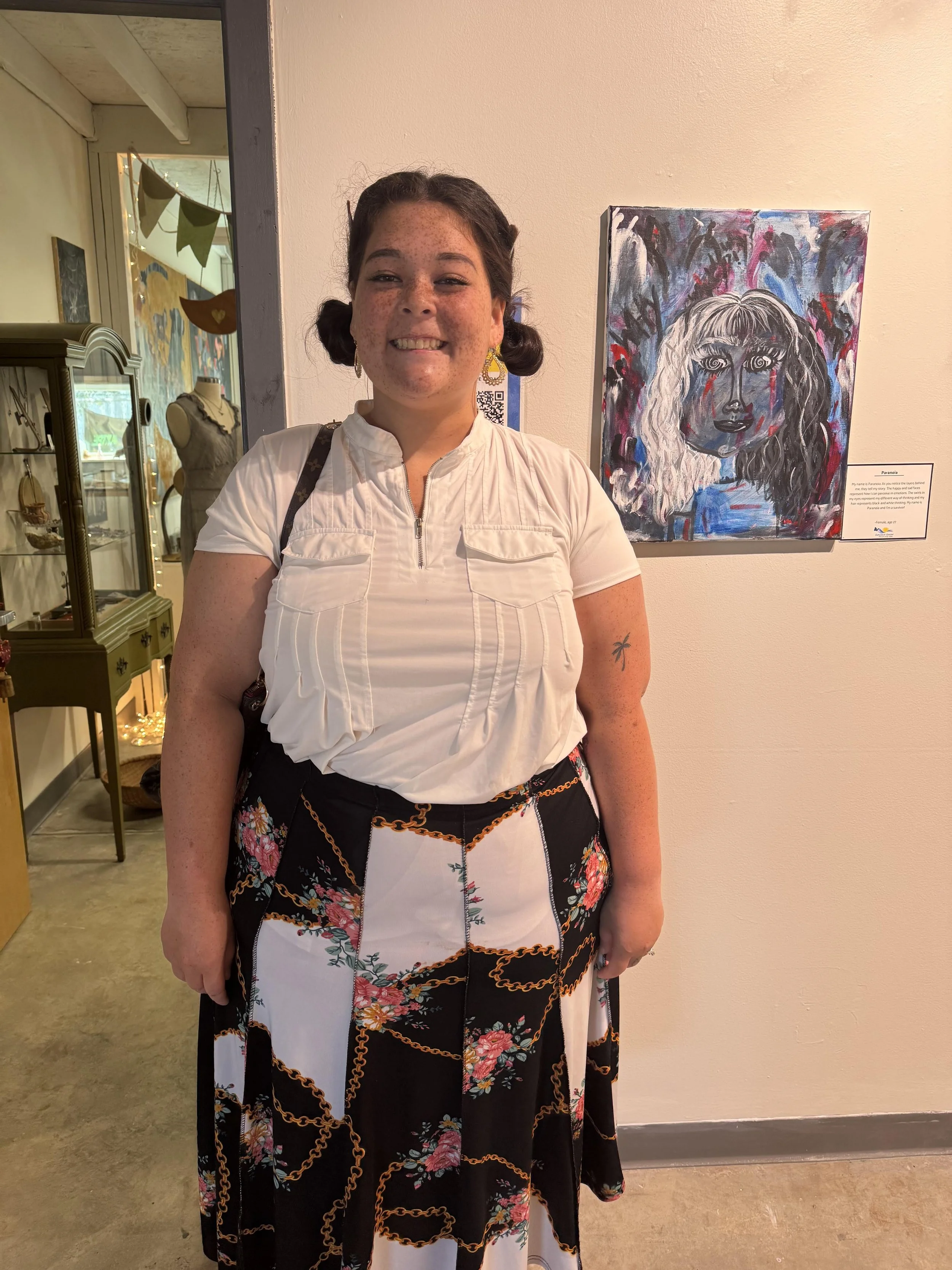 A woman smiling and standing in an art gallery. She has dark hair styled in pigtails, wears a white top and a long skirt with floral and chain patterns, and has a tattoo of a palm tree on her arm. Behind her, a colorful painting of a woman's face and a label with information about the artwork are visible.