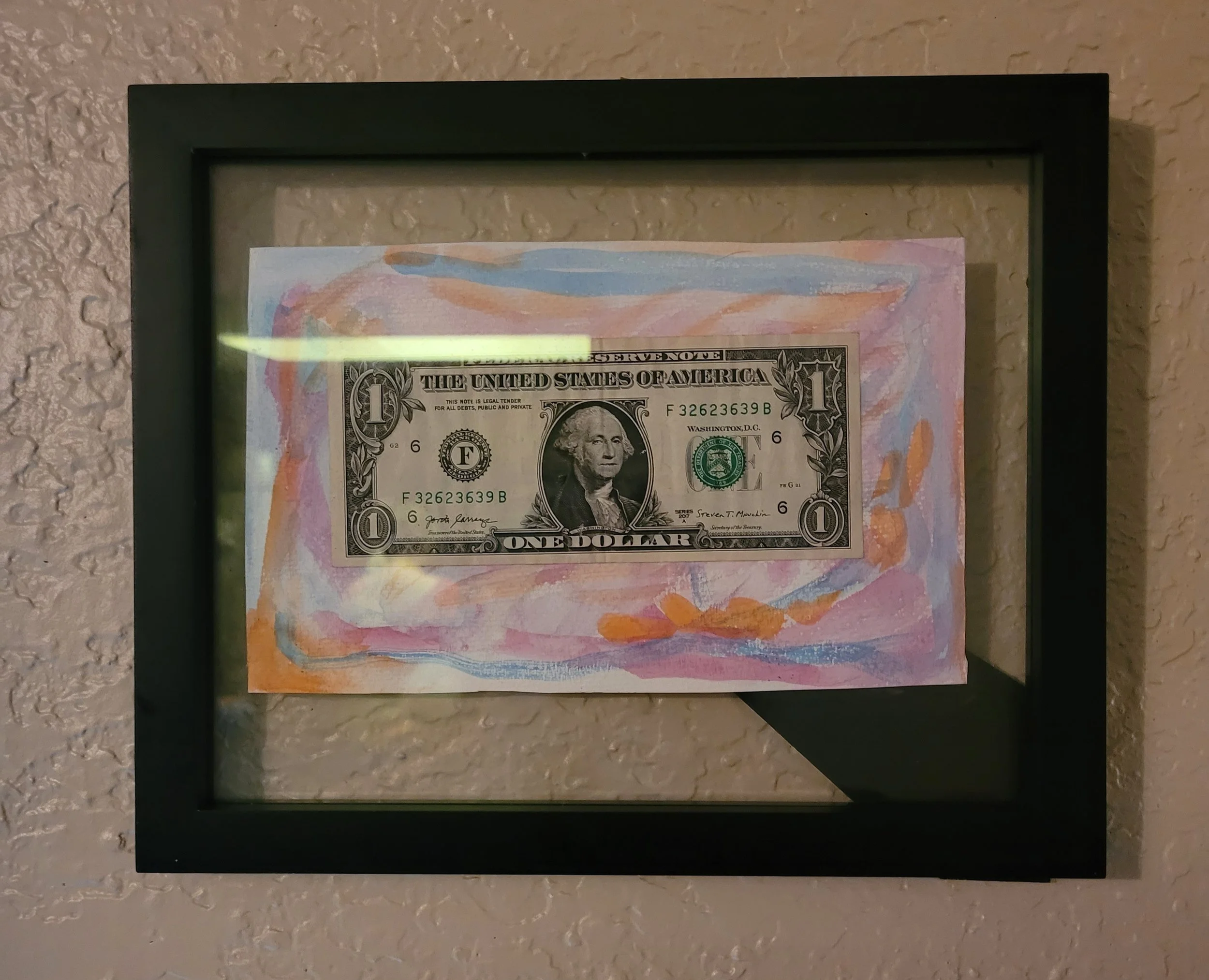 Framed artwork of a one-dollar bill on colorful abstract background, mounted on a textured wall.