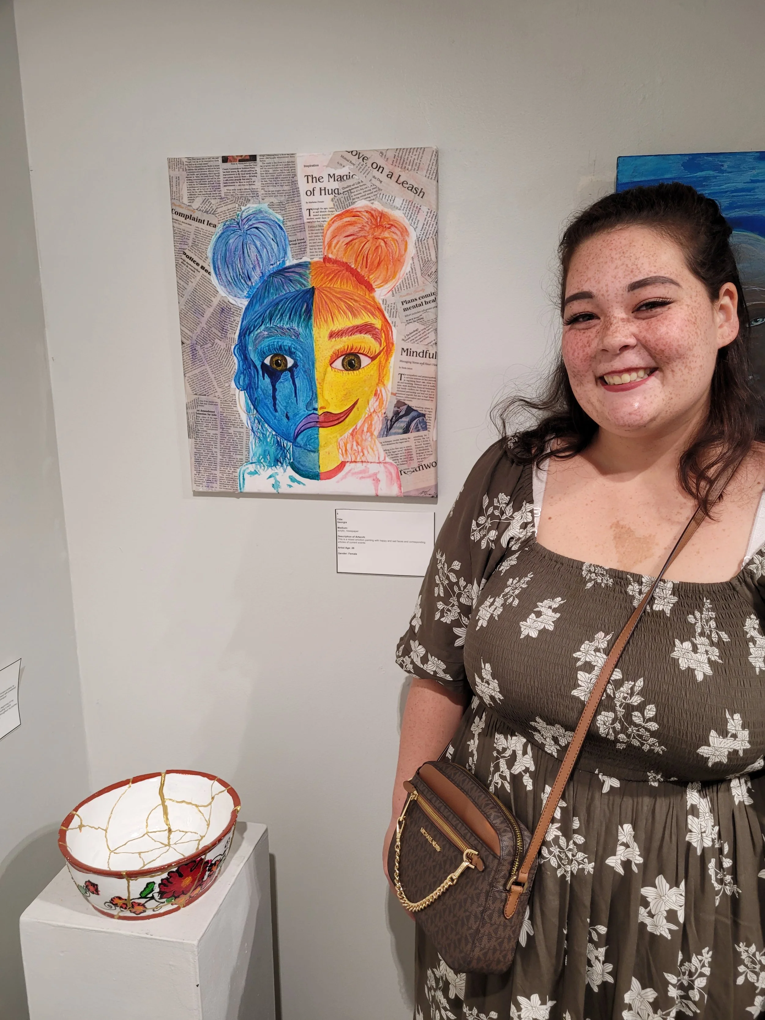 A woman with dark brown hair and freckles smiling at an art exhibit, standing next to a mixed media artwork of a girl's face divided into two halves: one side painted blue with tears, and the other side painted yellow, both set against a collage of newspaper clippings. A decorated cracked bowl is on a pedestal nearby.