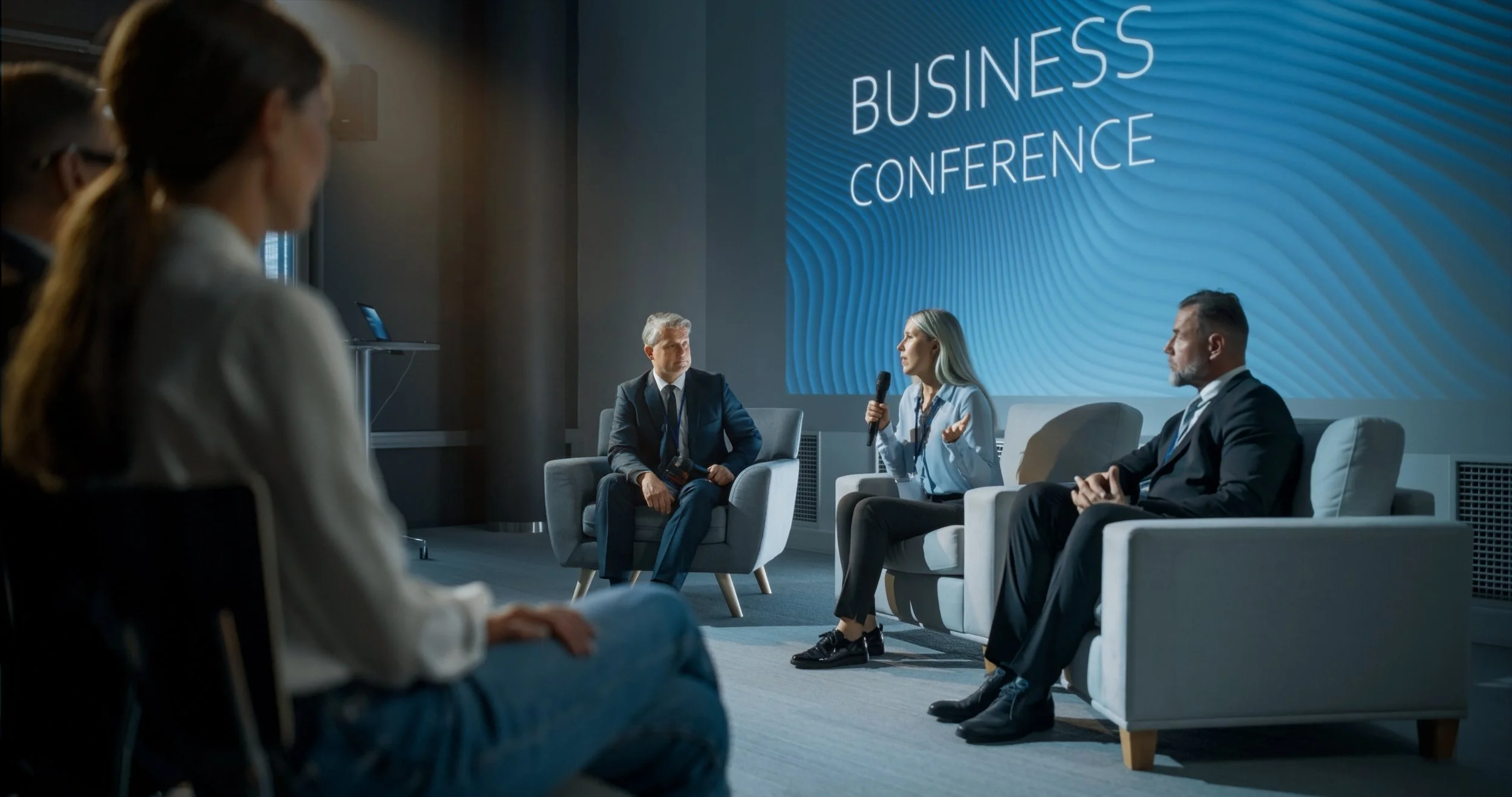 A panel discussion at a business conference, with four professionals sitting on stage chairs, one woman speaking into a handheld microphone, and a large screen behind. Sound reinforcement system for large audience.