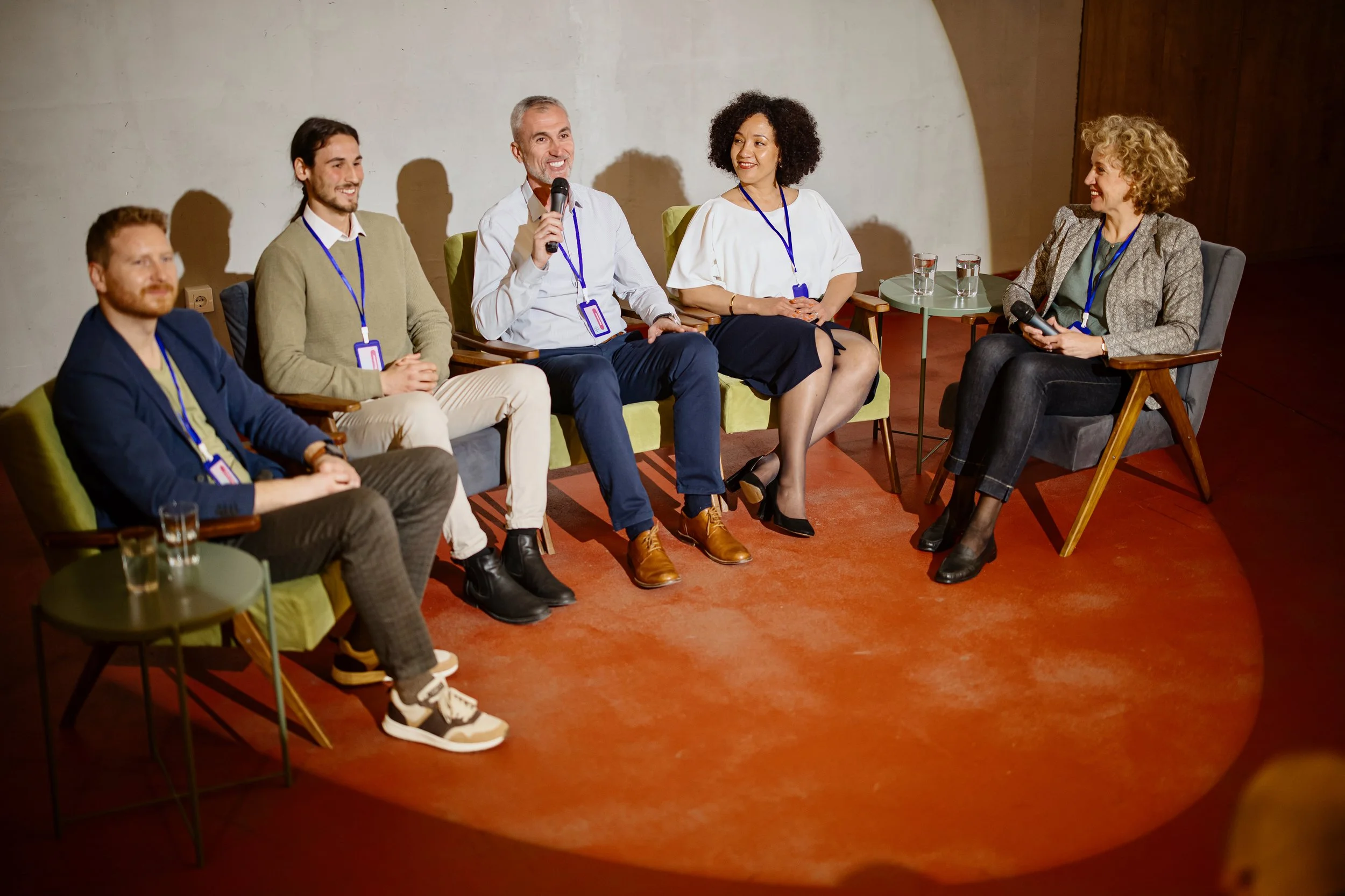 Group of six people sitting on chairs on stage during a panel discussion. One person in the middle is smiling and speaking into a handheld microphone. Spot lighting on the stage and full audio system reinforcement for medium audience.