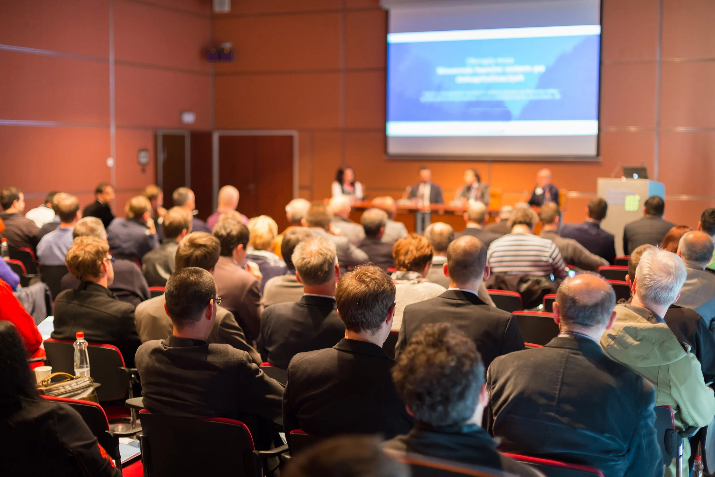 A conference room filled with attendees watching a panel discussion at the front, with a large screen displaying a presentation. Sound reinforcement for large venue.