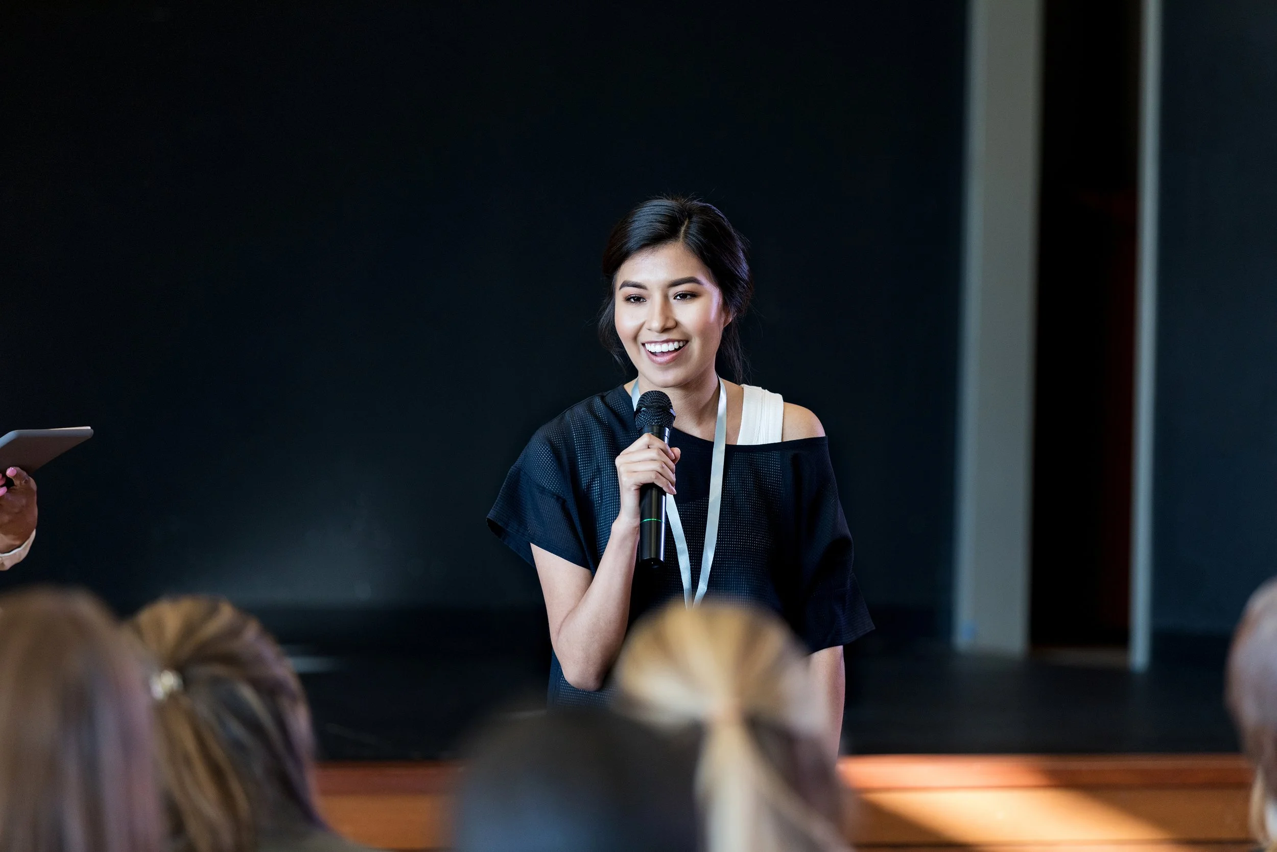 Young woman speaking into a microphone during a presentation or event, smiling with audience members visible in the foreground.