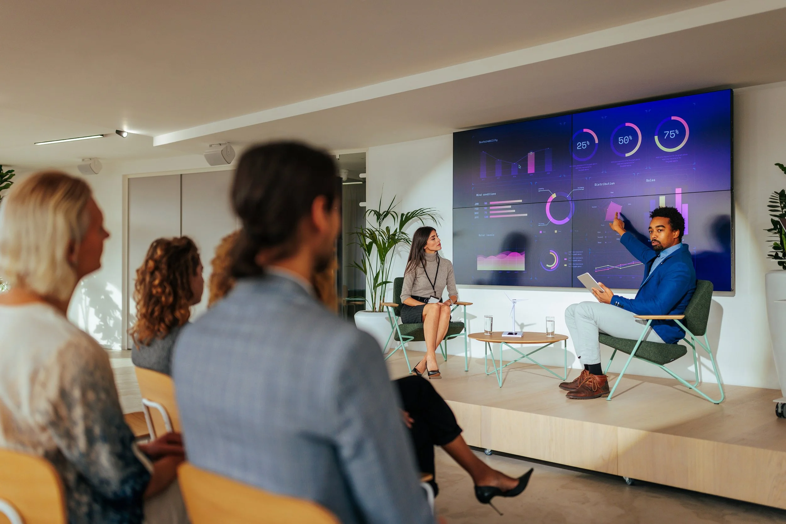 Business presentation in conference room with man in blue blazer explaining data on large digital screen to audience of diverse professionals.