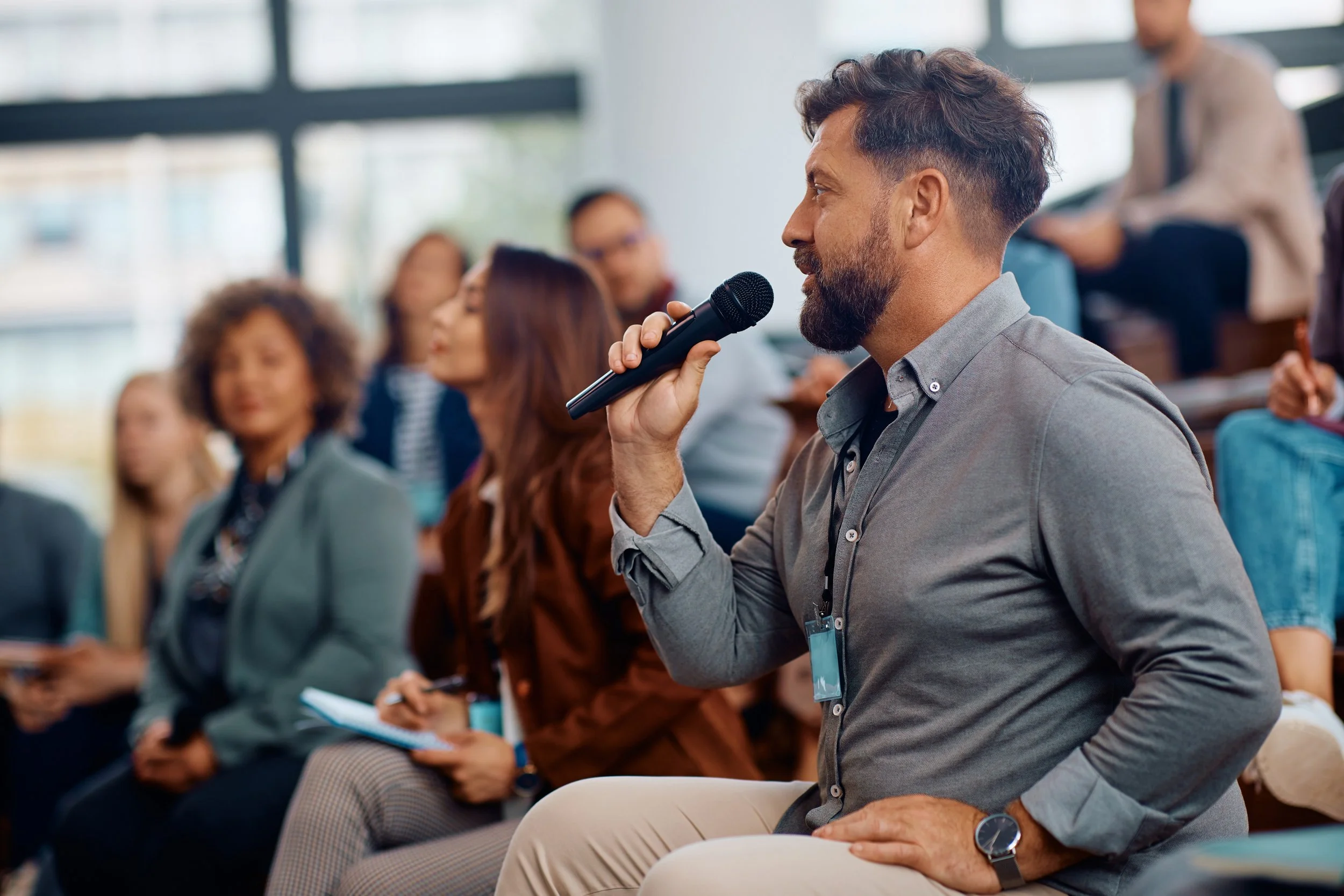 A man speaking into a microphone during a conference or seminar, surrounded by seated attendees, in a modern indoor setting.