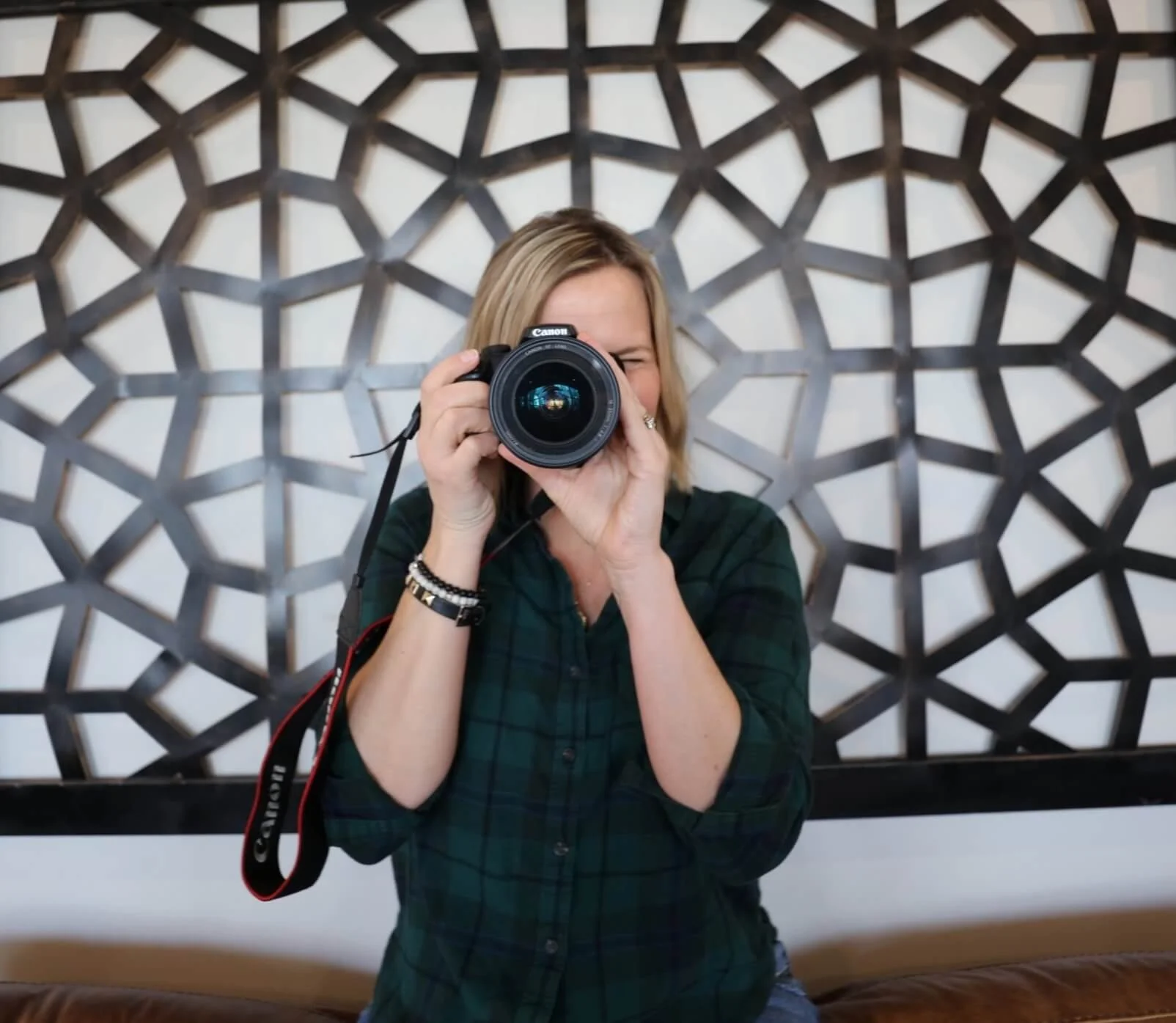 Person holding a camera and taking a photo, standing in front of a geometric patterned wall.