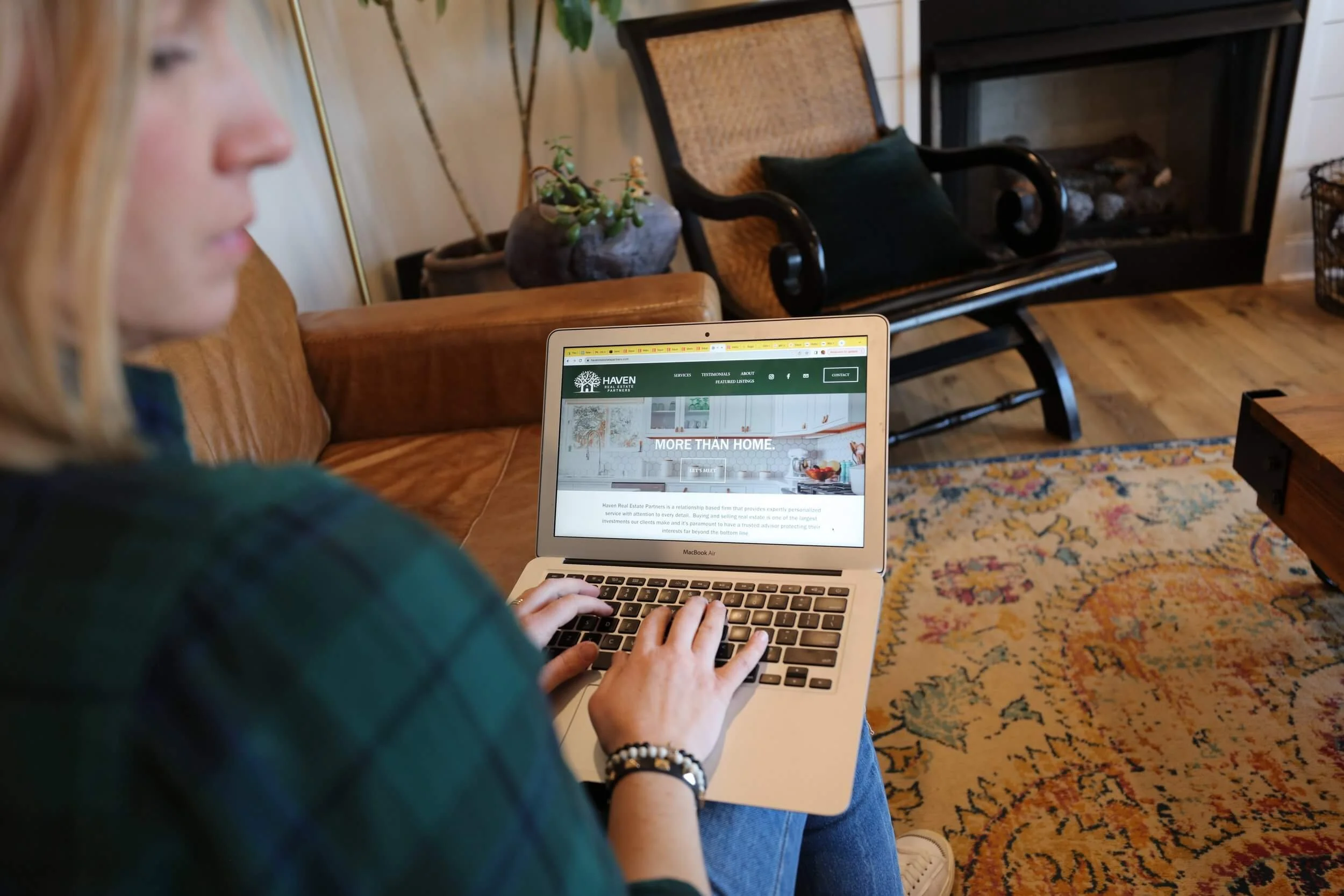 A woman sitting on a brown leather couch using a MacBook Air, viewing a real estate website titled 'Haven'. The room has wooden floors, a colorful rug, a black rocking chair with a cushion, a fireplace, and houseplants.