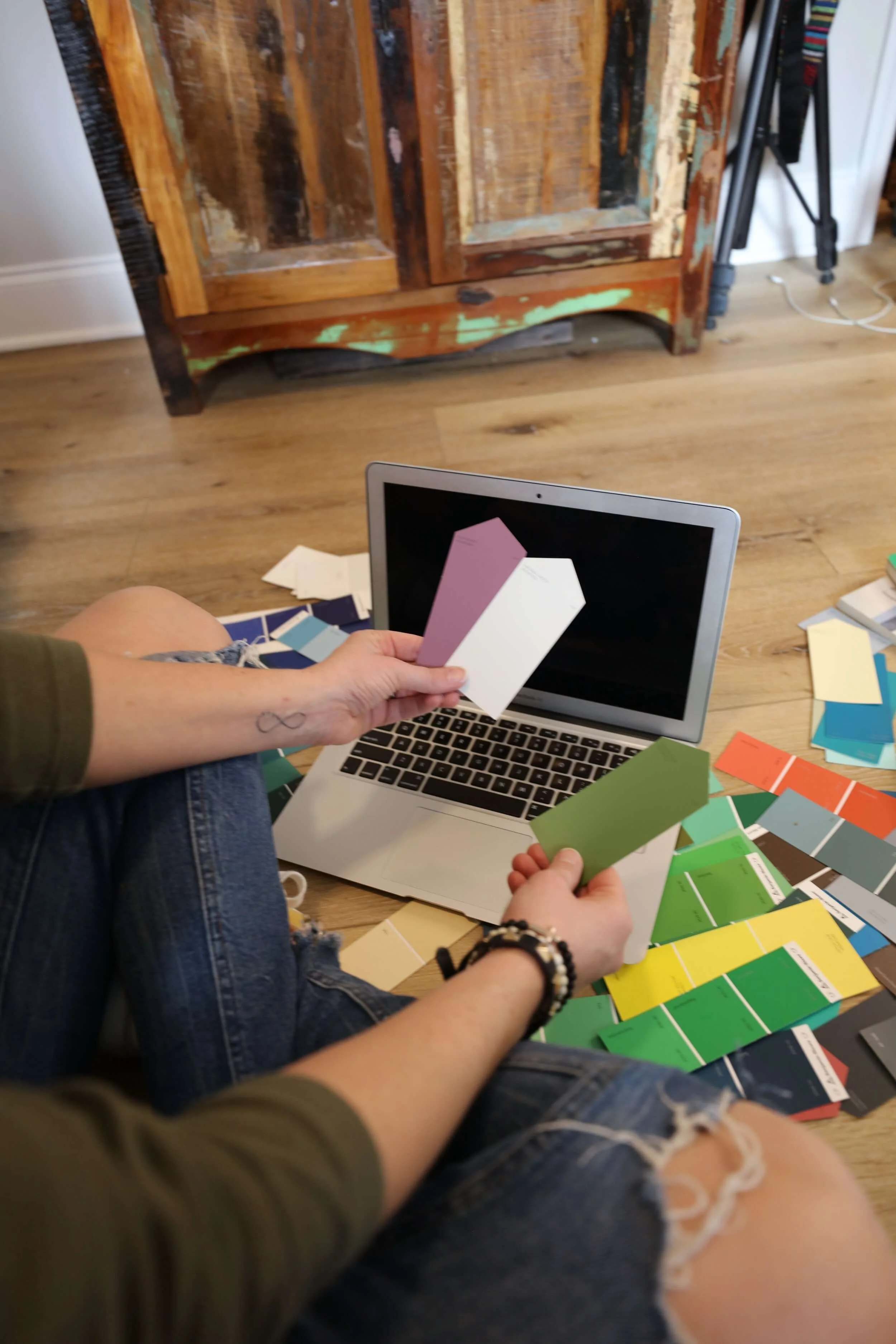 Person holding paint swatches in front of a laptop, surrounded by scattered color samples, wooden floor and rustic furniture in the background.