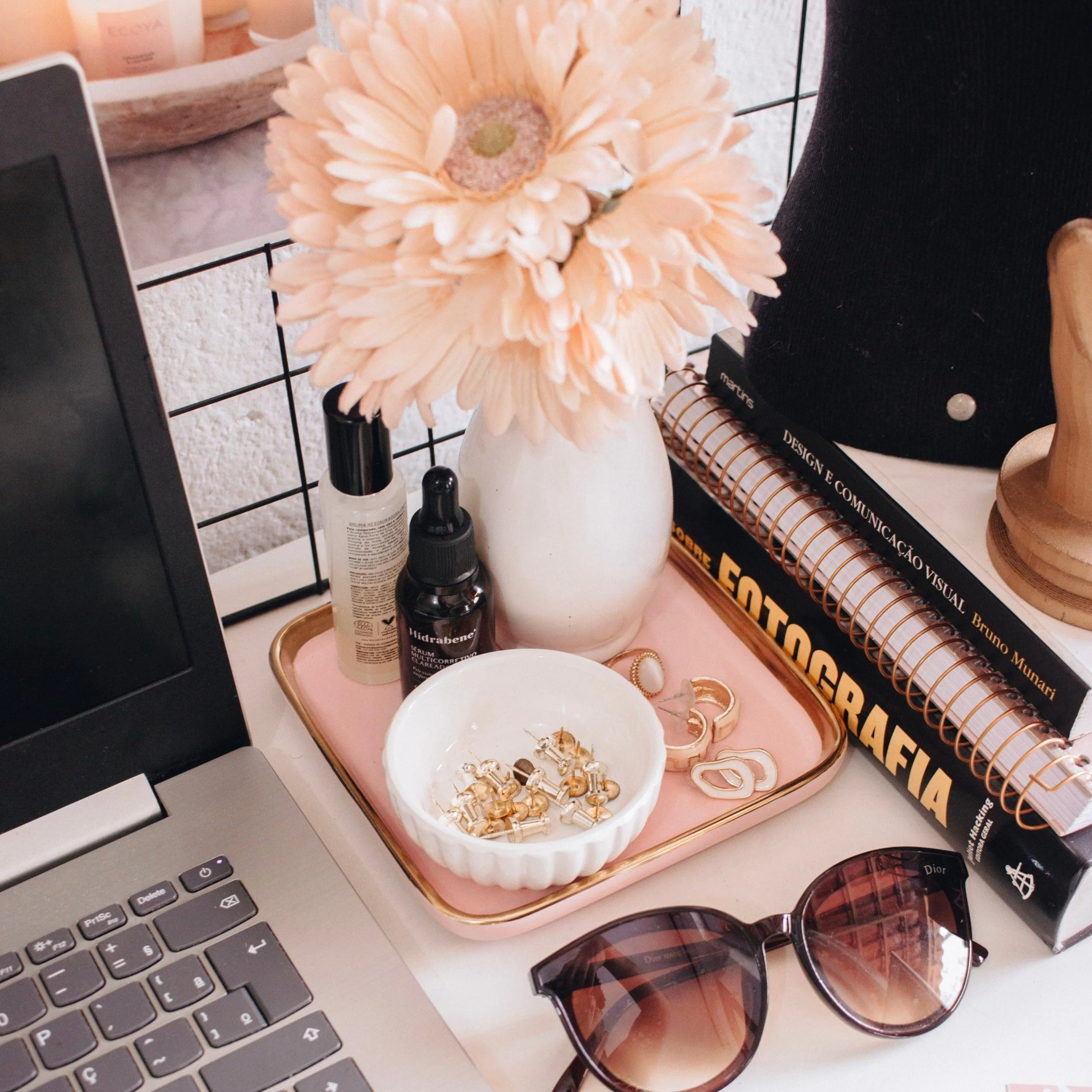 Desktop workspace with laptop, vase with flowers, sunglasses, skincare bottles, a bowl of jewelry, spiral notebooks, and a wooden ornament stand.