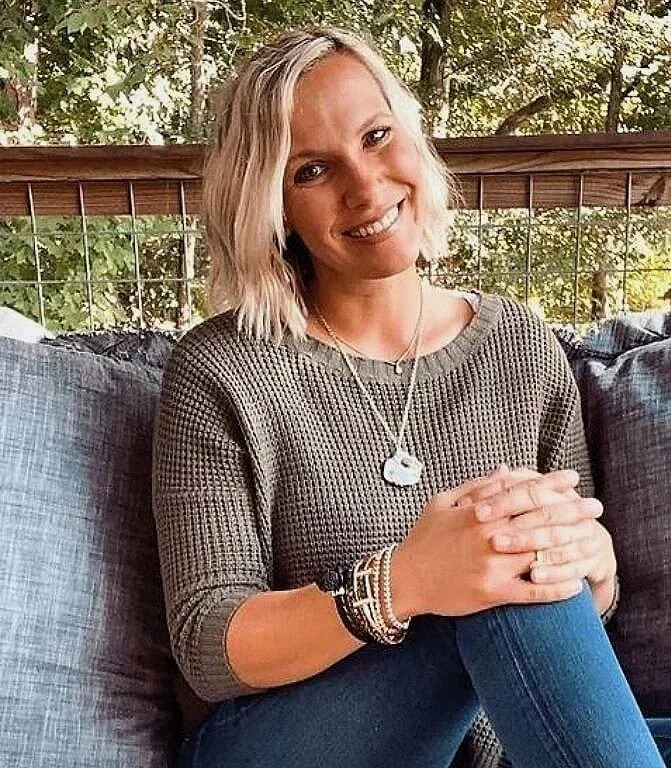 A woman with blonde hair sitting outside on a patio wearing a green sweater and blue jeans, smiling at the camera. Trees and a railing are visible in the background.