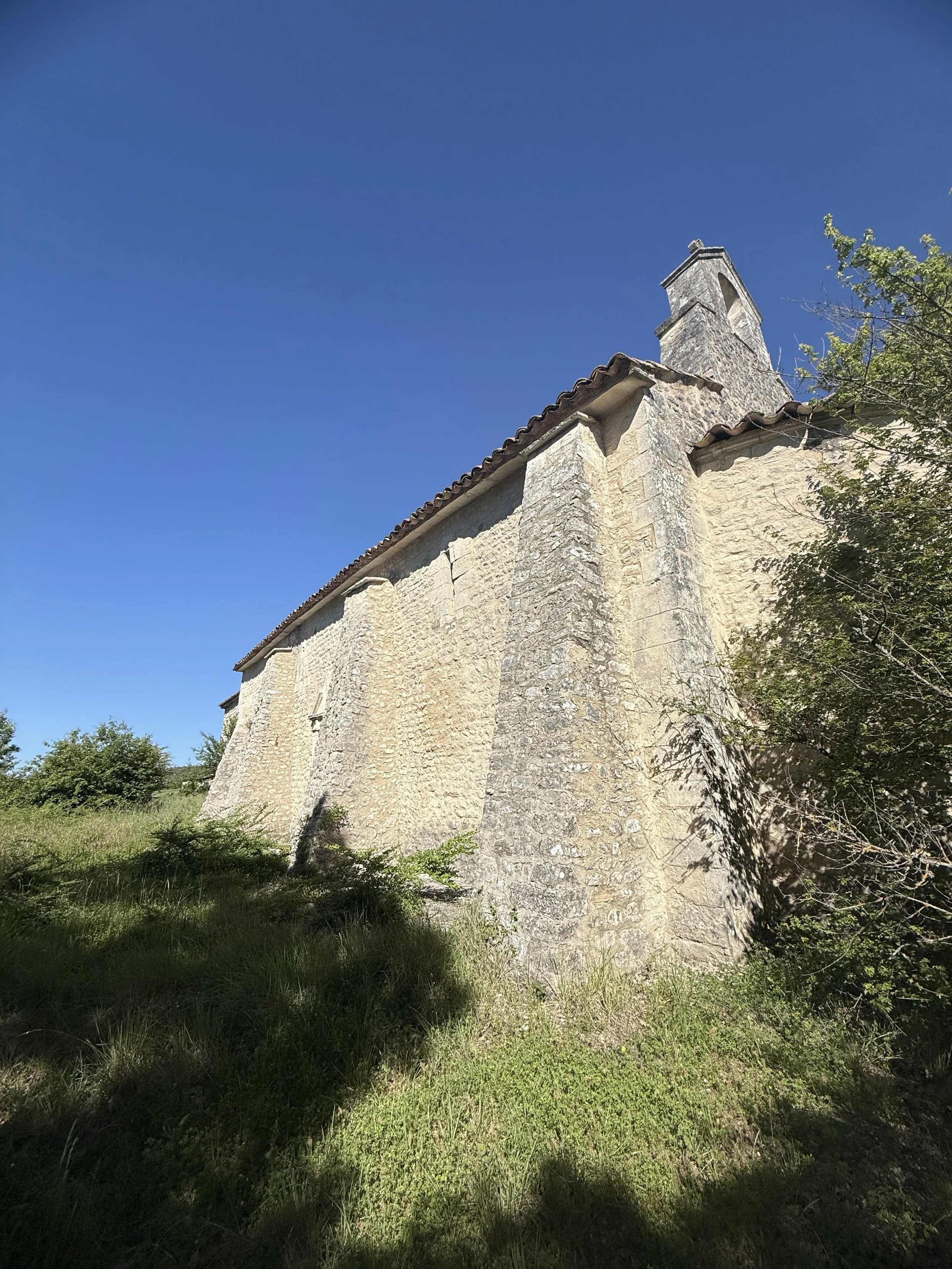 Vue d'une bâtisse en pierre avec un clocher, située sur une pente herbeuse, sous un ciel bleu clair.