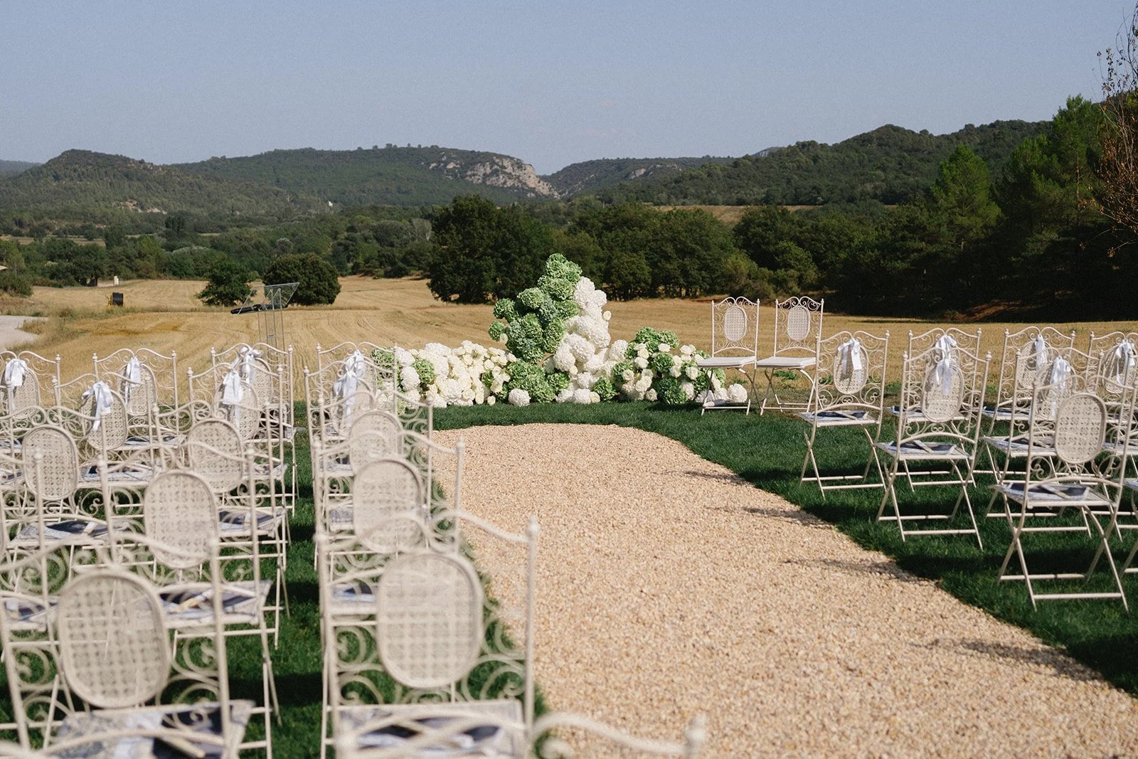 Allée de chaise blanches en fer forgé pour un mariage, avec décoration florale blanche et verte en fond, en plein air avec vue sur un paysage rural et des collines en arrière-plan.