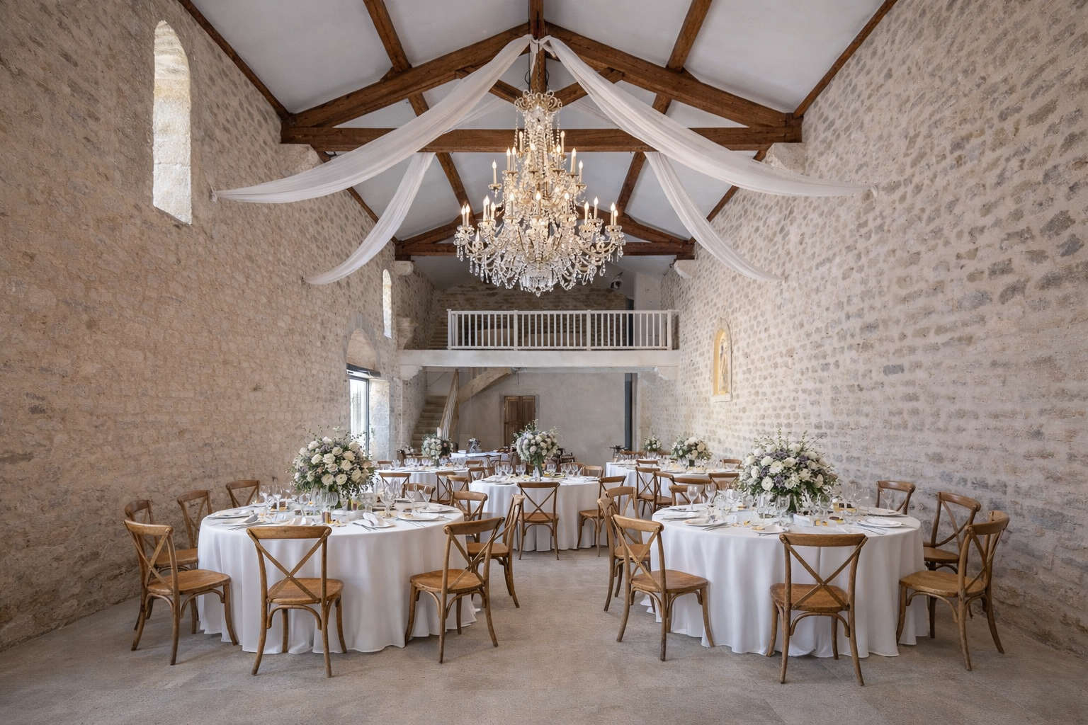 Salle de réception avec tables rondes ornées de nappes blanches, décorées de bouquets de fleurs blanches, dans une pièce avec murs en pierre et un chandelier en cristal au plafond.