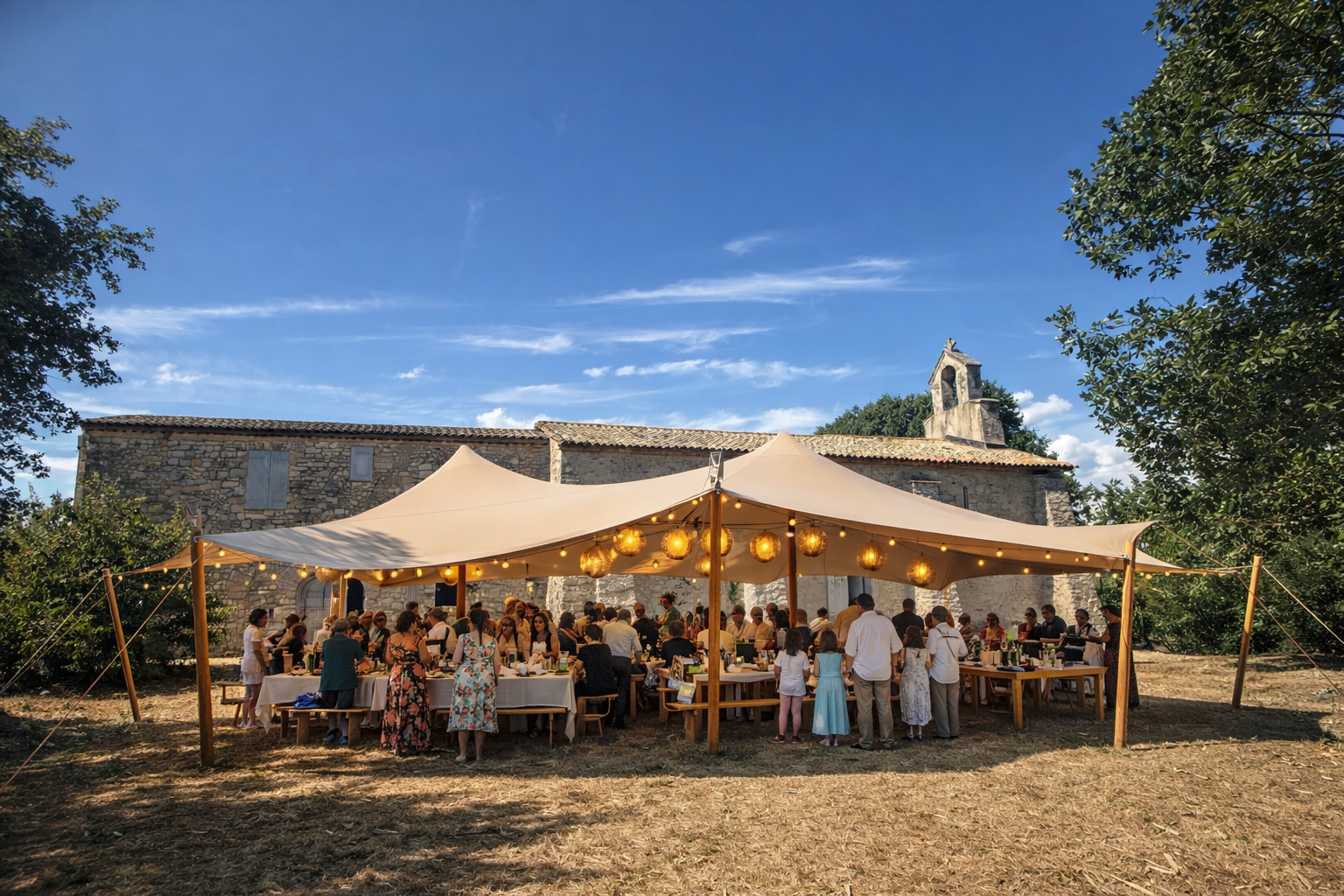 Une fête en plein air sous une tente beige décorée de lanternes, avec beaucoup de personnes autour de tables, dans un contexte rural avec un vieux bâtiment en pierre et une petite tour en arrière-plan, par un jour ensoleillé.