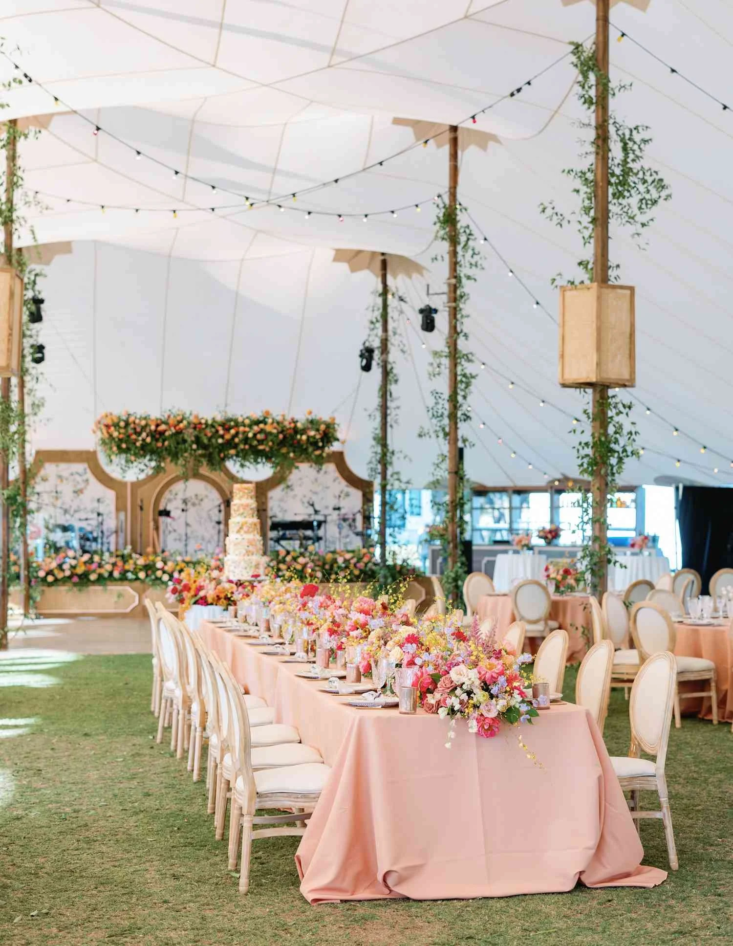 Une décoration de mariage avec une grande table recouverte d'une nappe rose, ornée de bouquets de fleurs colorées. Au fond, un autel décoré de fleurs et un gâteau de mariage. Le tout sous une grande tente avec des guirlandes lumineuses.