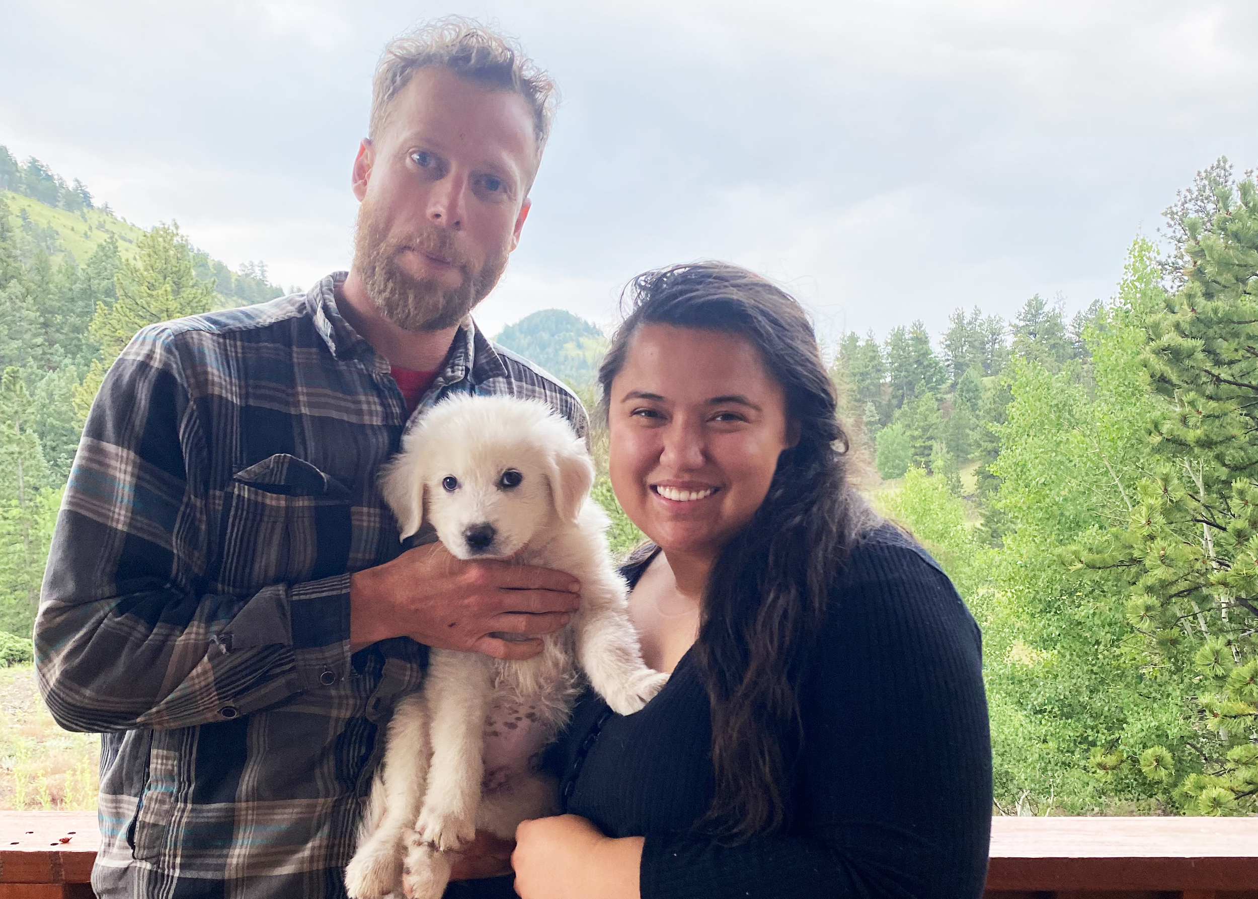 A man and woman smiling outdoors with a puppy, surrounded by trees and mountains.
