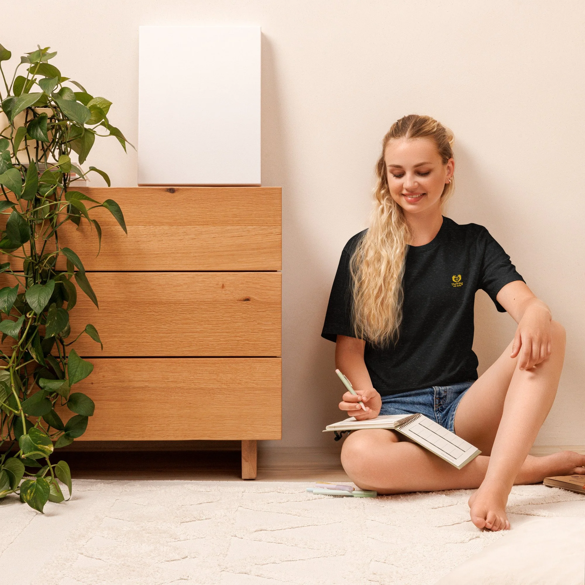 A young woman with long blonde hair, wearing a black T-shirt and denim shorts, sits on the floor smiling while writing in a notebook. She is next to a wooden dresser and a large green houseplant, with a white wall behind her.