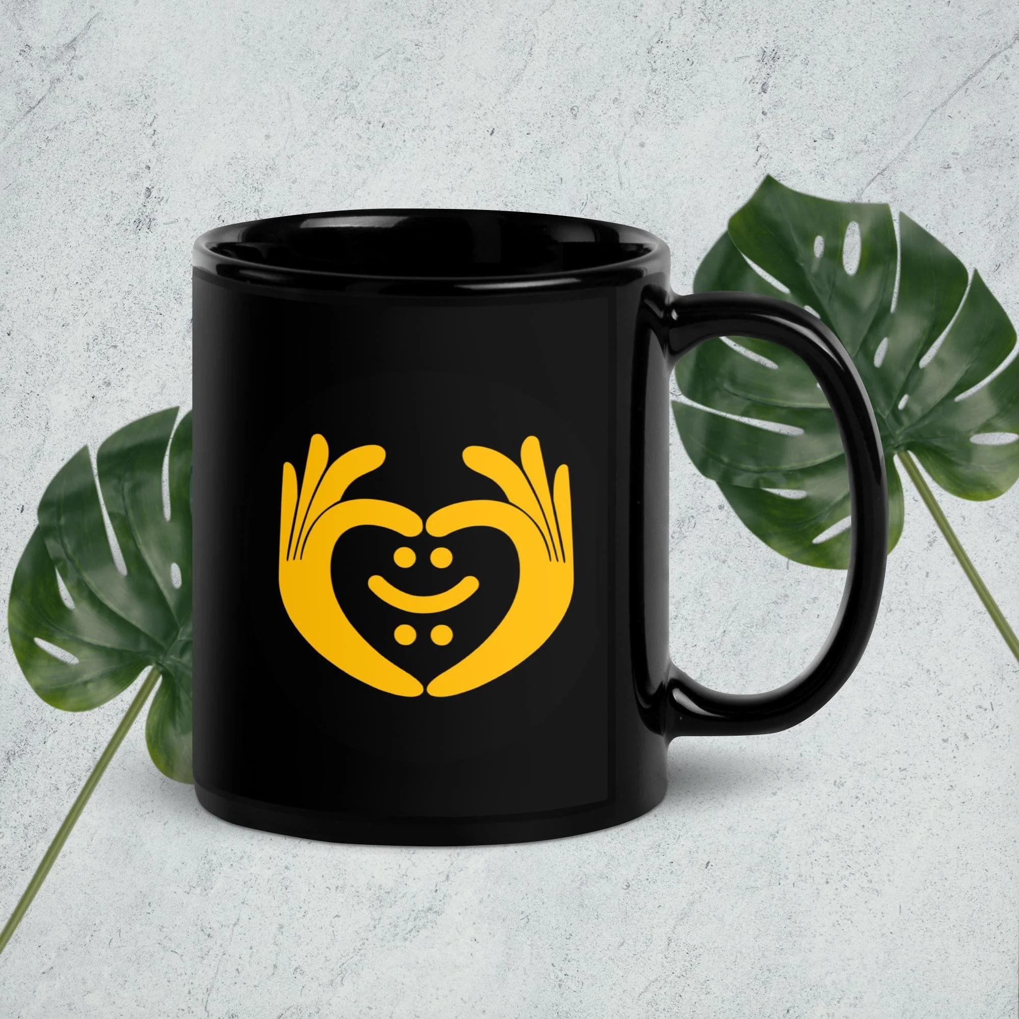 Black mug with yellow logo of two hands forming a heart shape around a face with eyes, nose, and mouth, placed on a light-colored textured surface with green monstera leaves in the background.