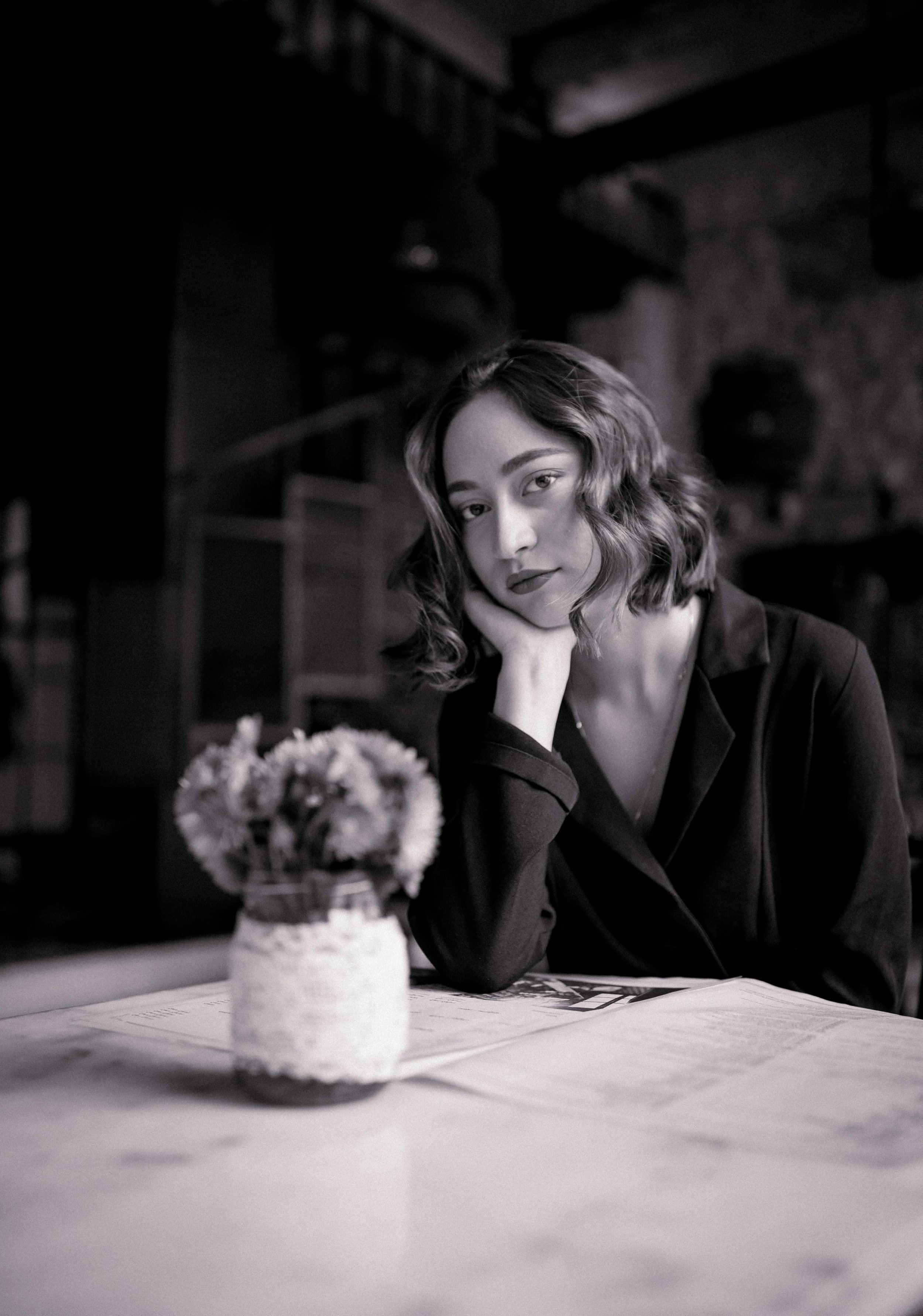 A woman with wavy hair sitting at a table, resting her head on her hand, with a vase of flowers and a newspaper in front of her. The photo is in black and white.