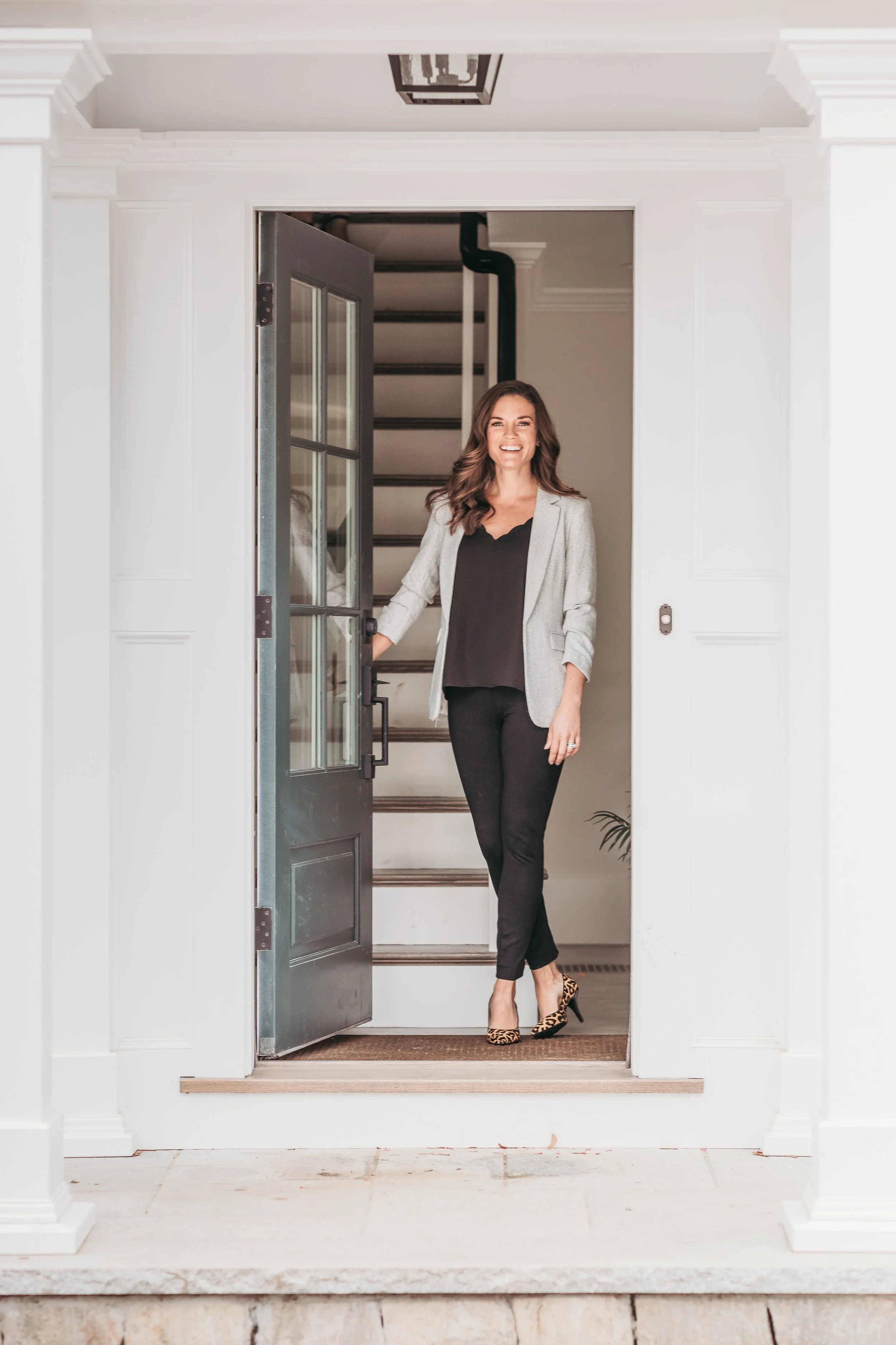 A woman stands at the doorway of a house, smiling, wearing a gray blazer, black top, black pants, and leopard-print high heels.
