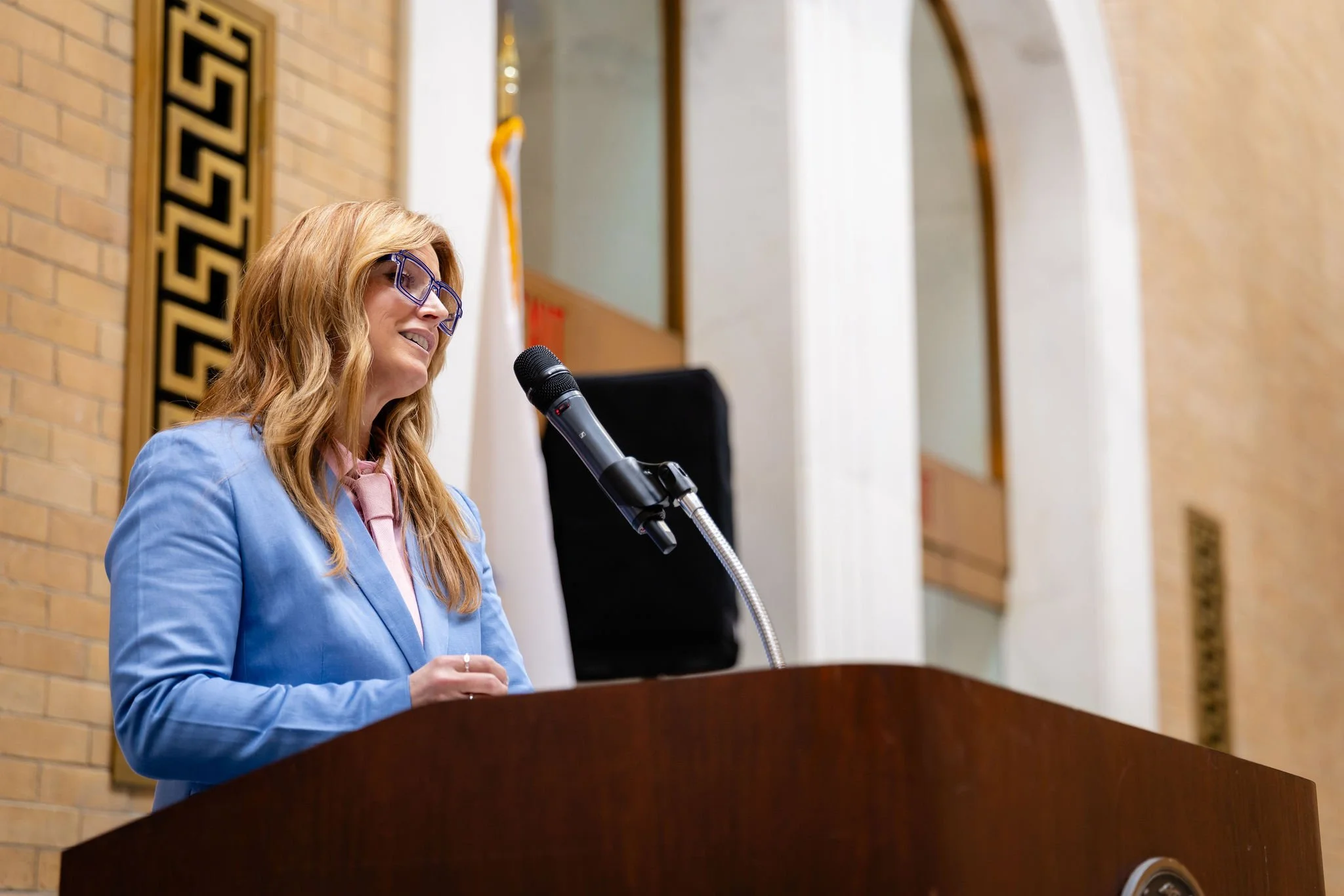 A woman with long blonde hair in a blue blazer, pink blouse, and purple eyeglasses speaking into a microphone at a podium in a room with brick walls and large windows.