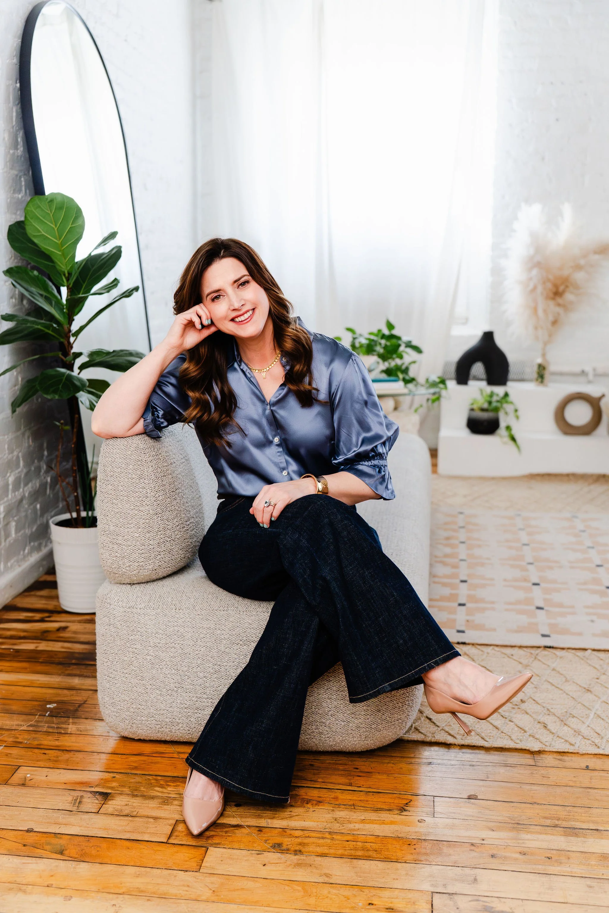 A woman sitting on a beige armchair in a bright room with white brick walls, plants, and decorative objects in the background. She has long brown hair, is smiling, wearing a shiny blue blouse, dark jeans, and nude high heels.
