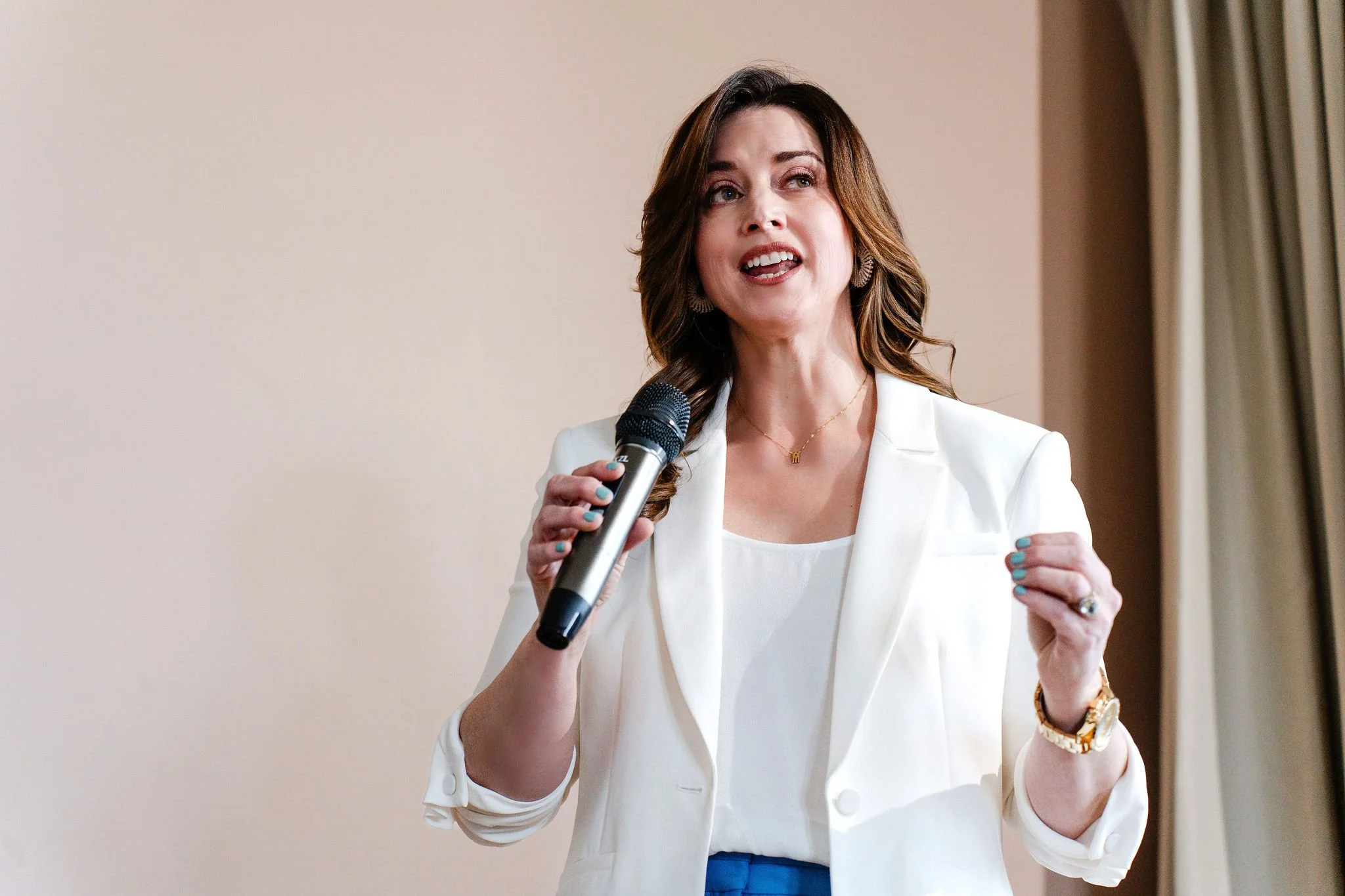 A woman with brown hair and earrings wearing a white blazer and blue pants speaking into a microphone.