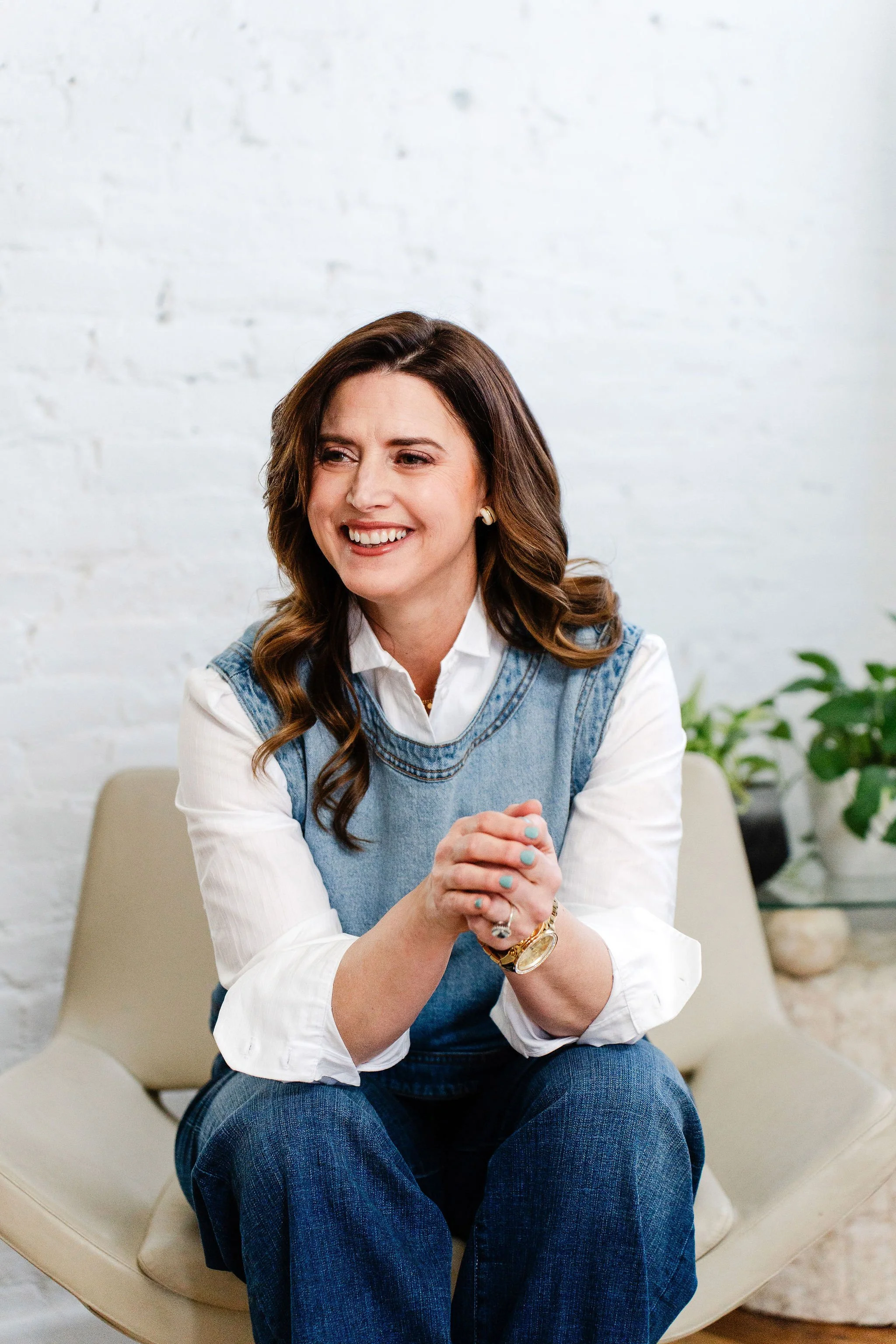 A woman with long, wavy brown hair, smiling, wearing a white shirt and a denim vest, sitting on a beige chair with crossed hands, in a room with white brick wall and green plants.