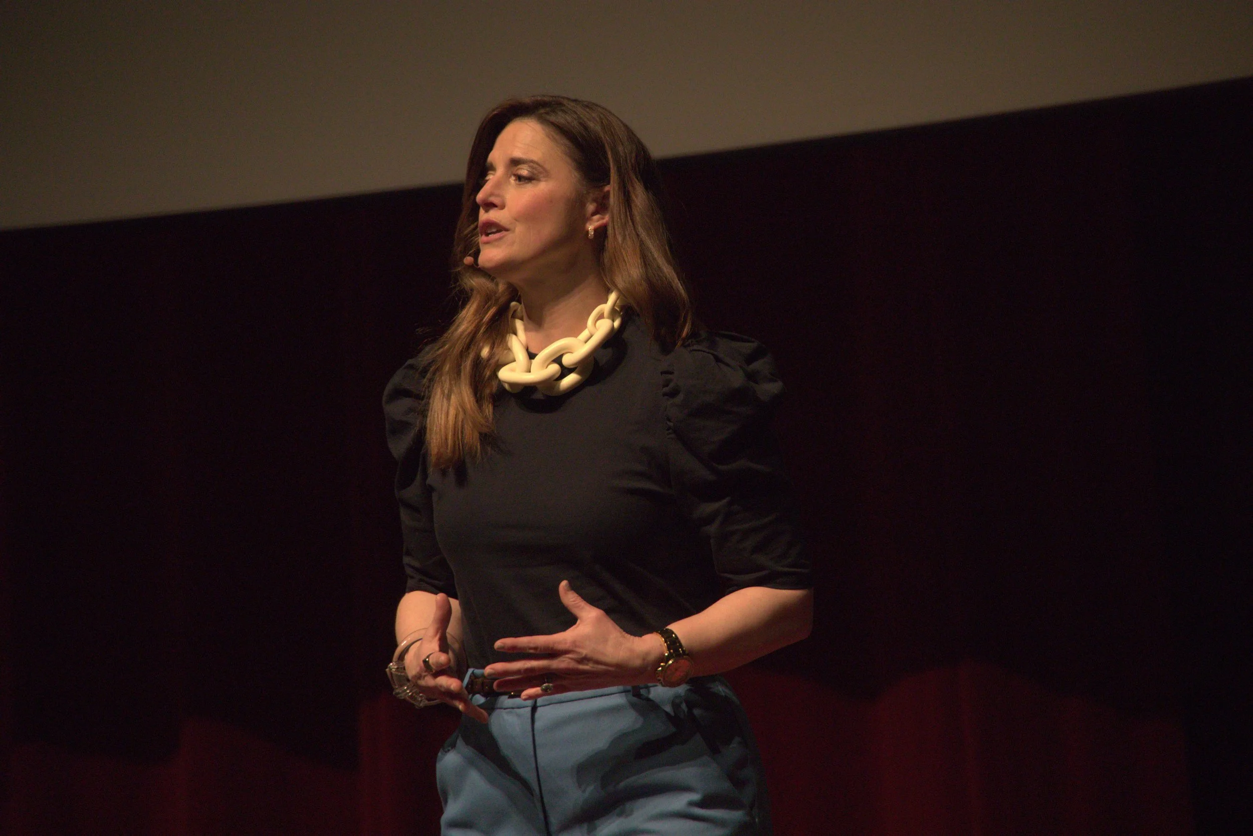A woman with long brown hair wearing a black outfit and a large white statement necklace speaking on stage.
