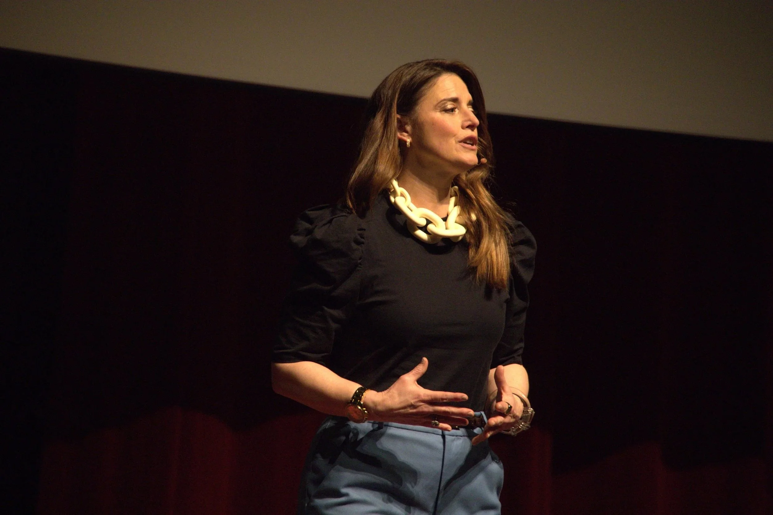 Woman speaking on stage, wearing black dress and chunky white necklace, with her hand on her hip.