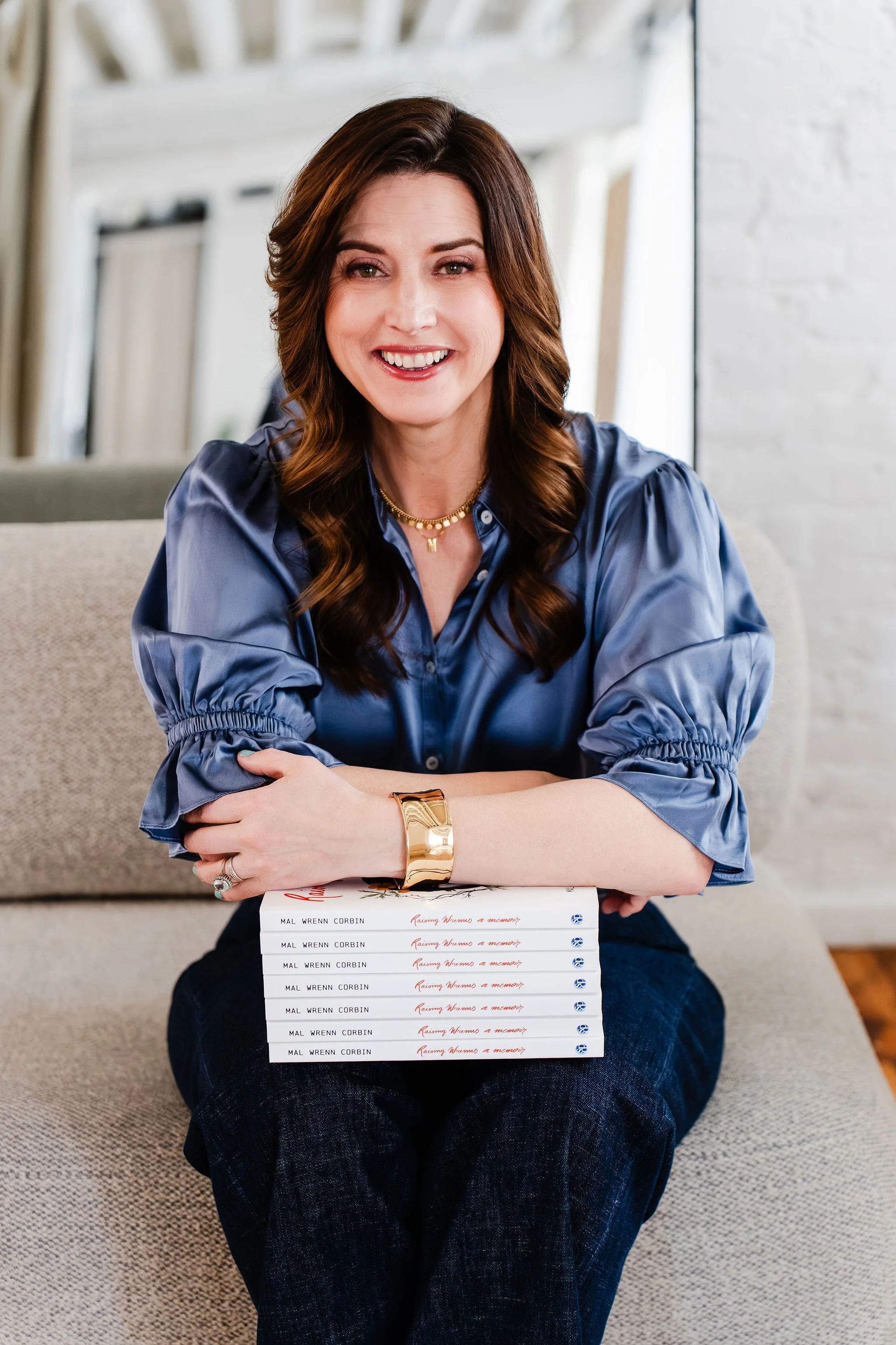 A woman with wavy brown hair, wearing a blue satin blouse, is smiling at the camera. She is sitting on the floor with a stack of books titled "Raising Wires a Memory" by Mal Wrenn Corbin on her lap. The background shows a cozy indoor setting with a light-colored sofa and white walls.
