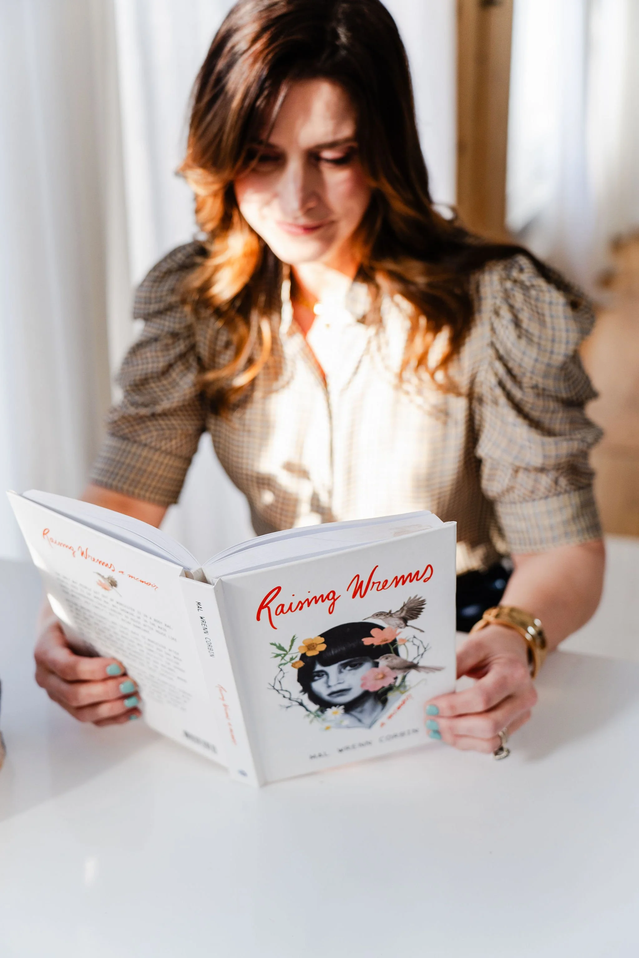 A woman with long brown hair reading a book titled 'Raising Wremms' while sitting at a white table, with a blurred background of a room with curtains.